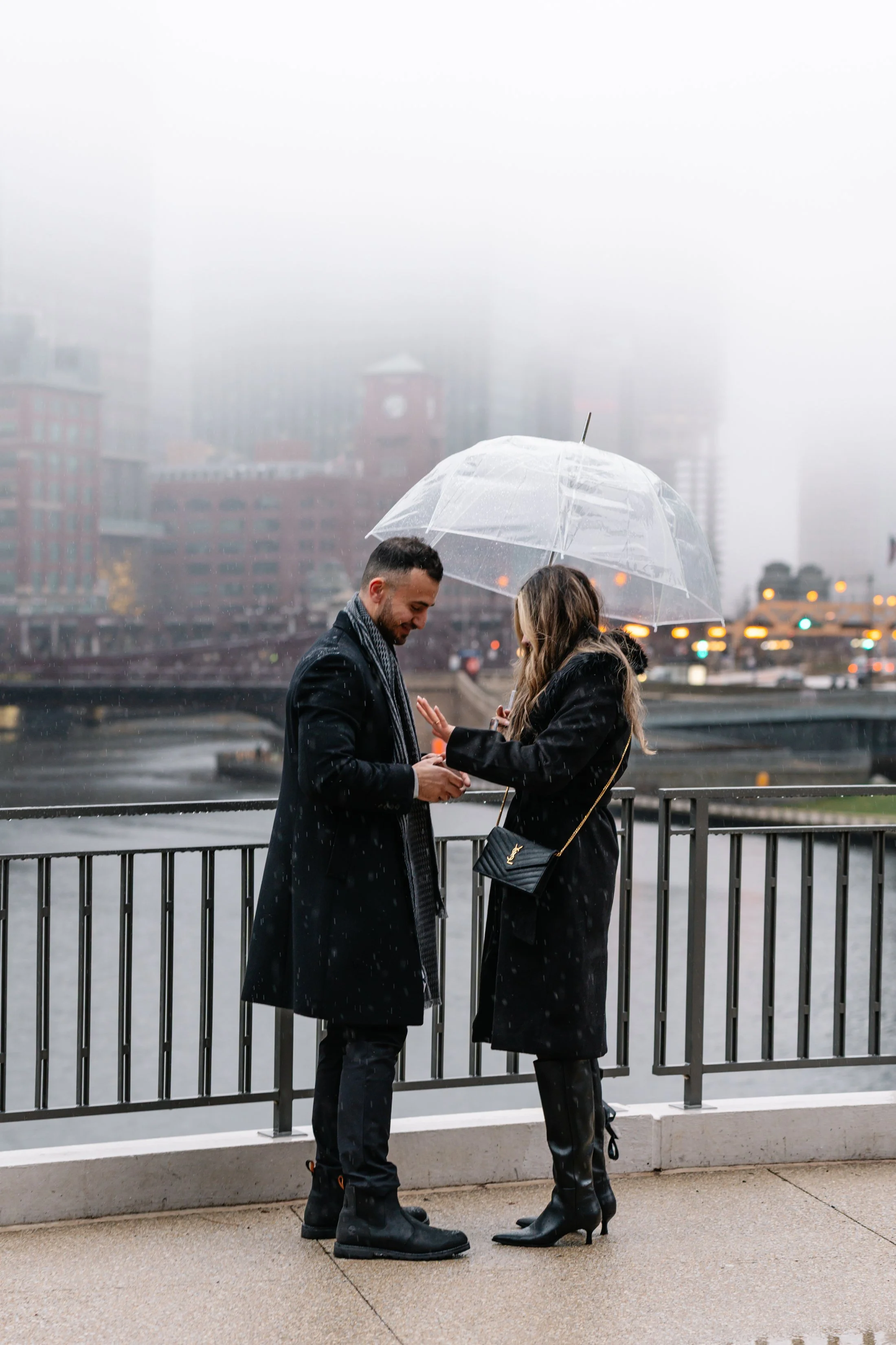 A man and woman share a moment under a transparent umbrella on a rainy day, with city buildings and water in the background.