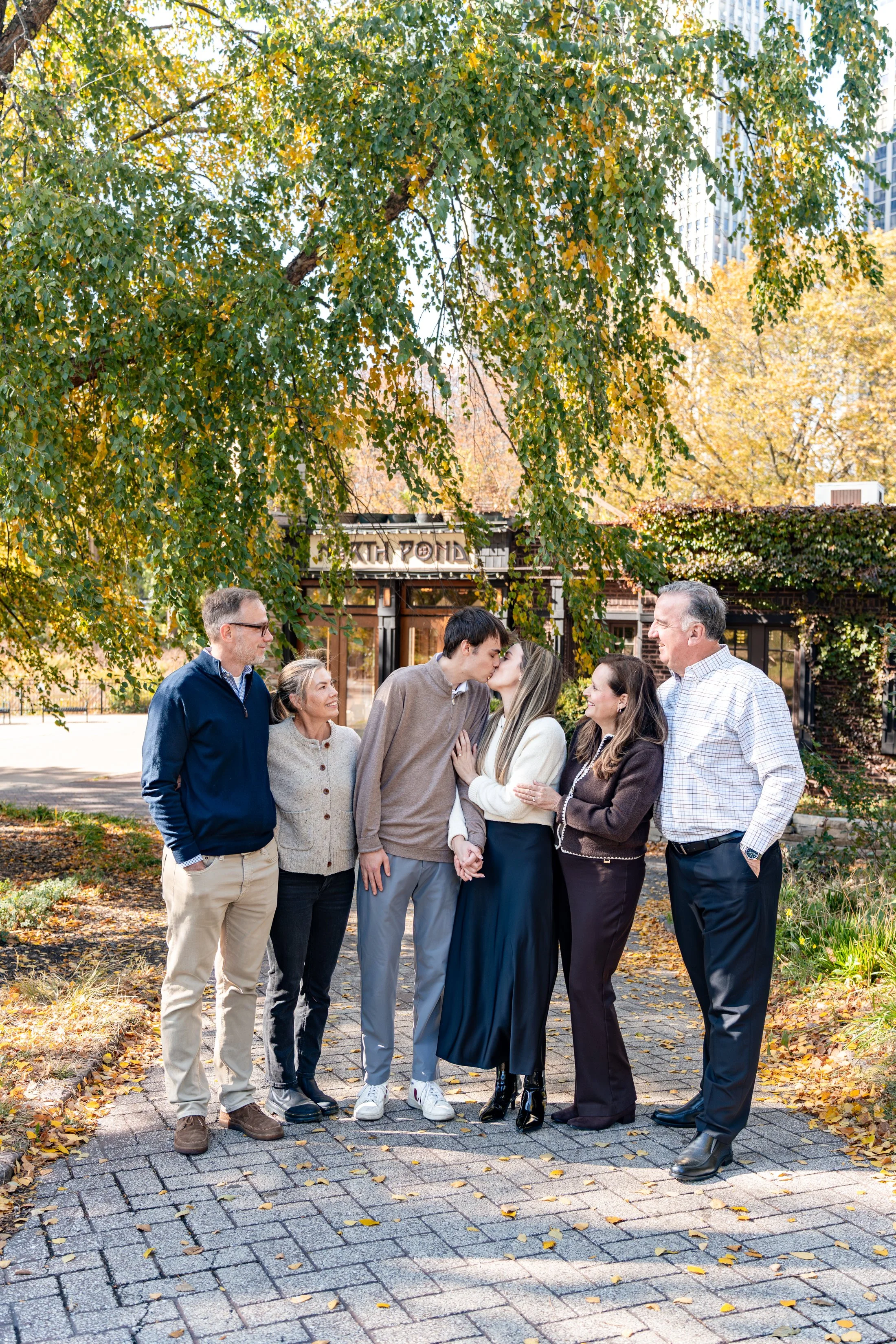 Family of six standing outdoors under a large tree, with two young adults kissing and the others smiling and talking, in front of a building with a sign that says 'South Point' and autumn foliage.