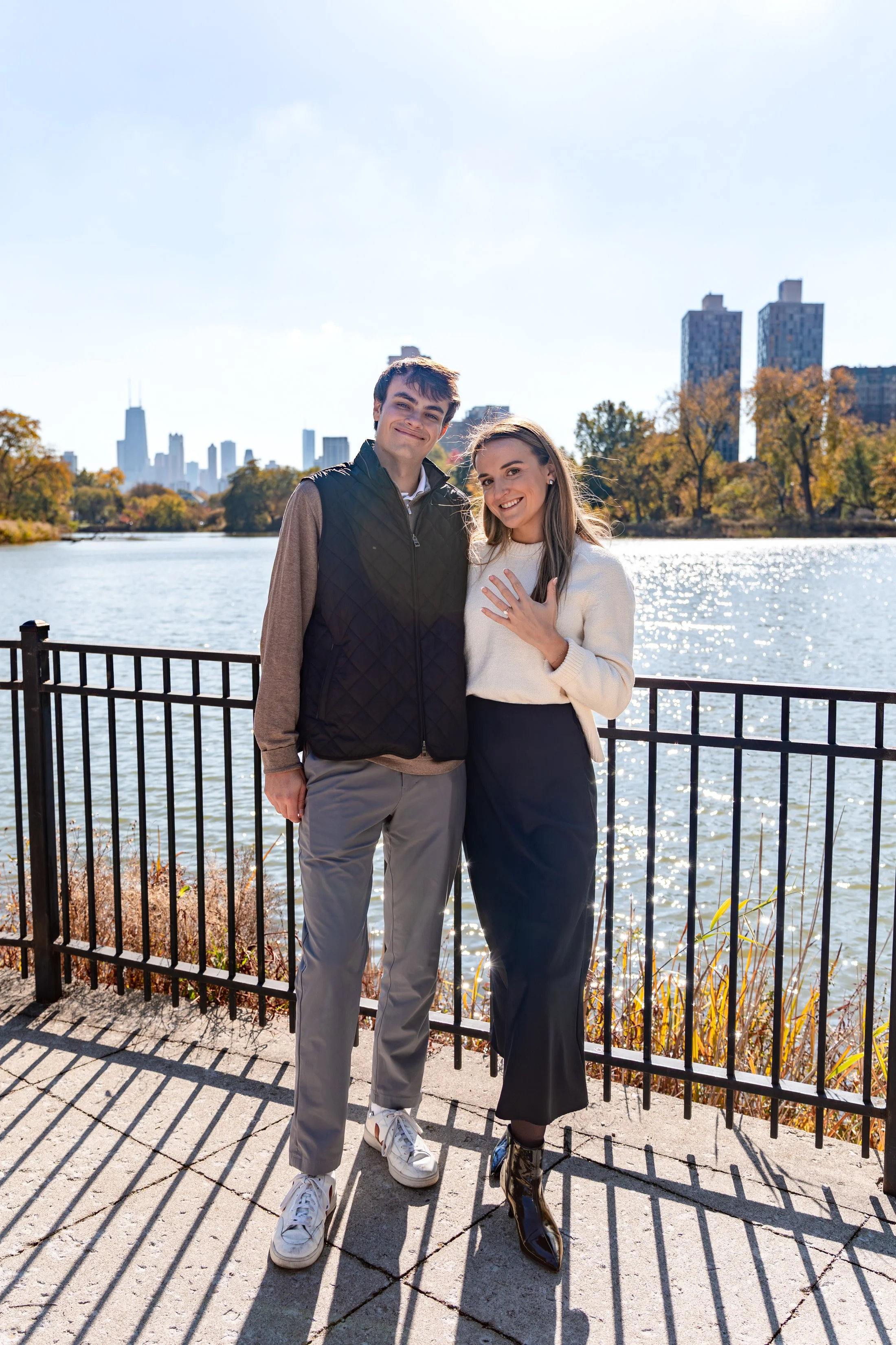 A young couple standing by a lake with a city skyline in the background during autumn, smiling and showing their rings.