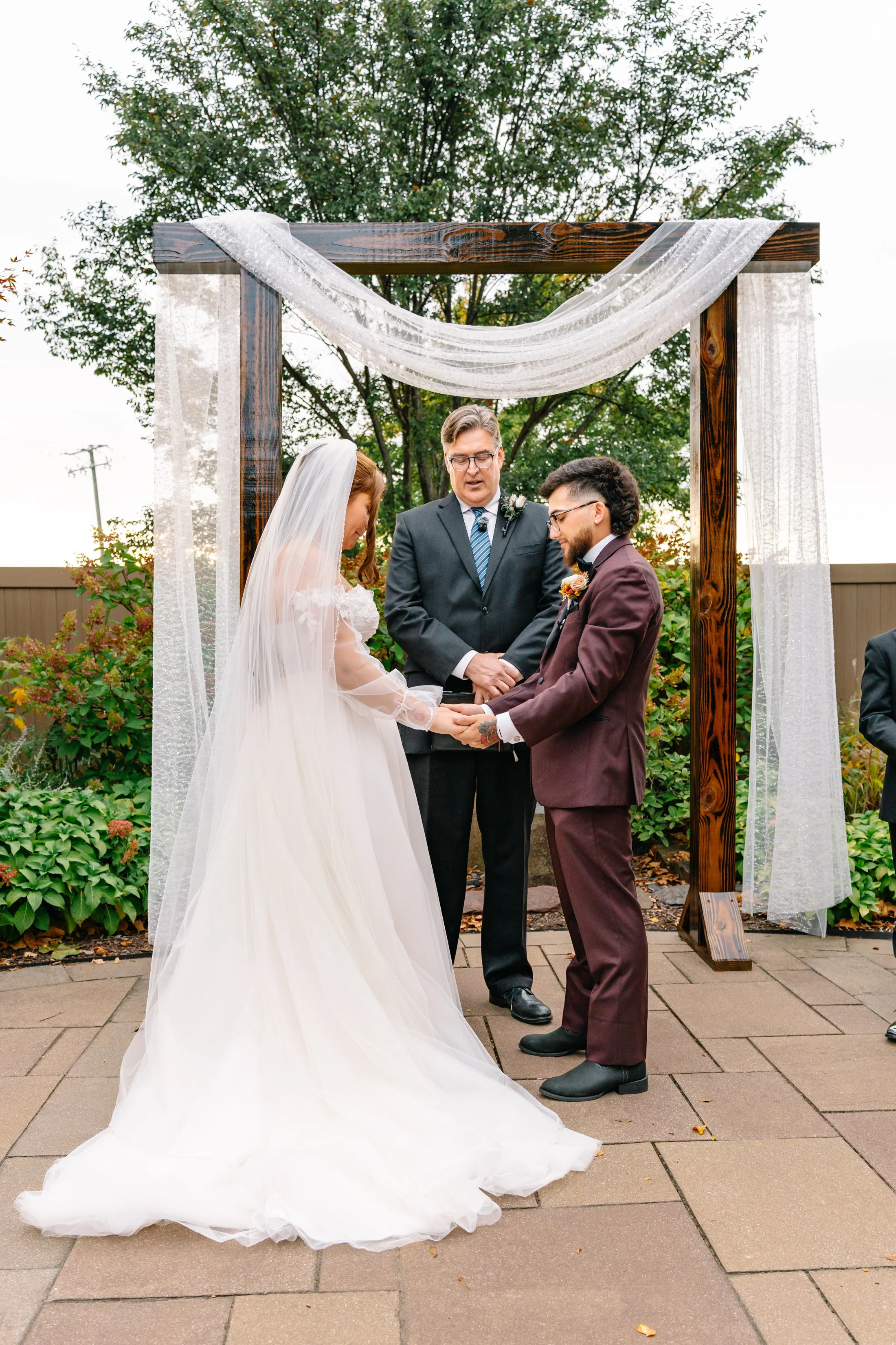 A couple getting married outdoors under a wooden arch decorated with white curtains, with an officiant standing between them.