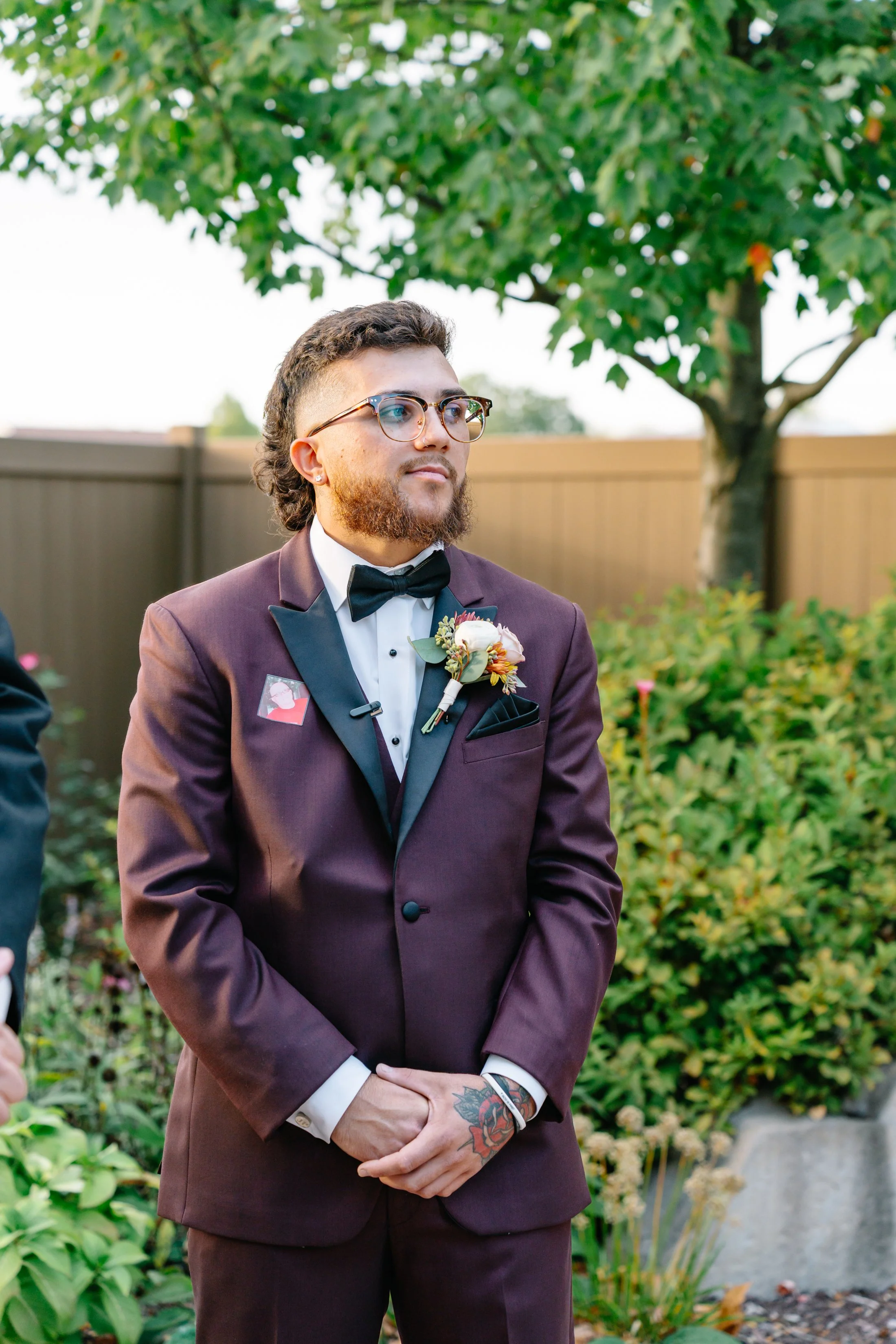 A man dressed in a burgundy tuxedo with a black satin lapel, white shirt, and black bow tie standing outdoors in a garden setting. He has glasses, a boutonniere on his left lapel, and a tattoo on his left wrist. Behind him are green shrubs, trees, an