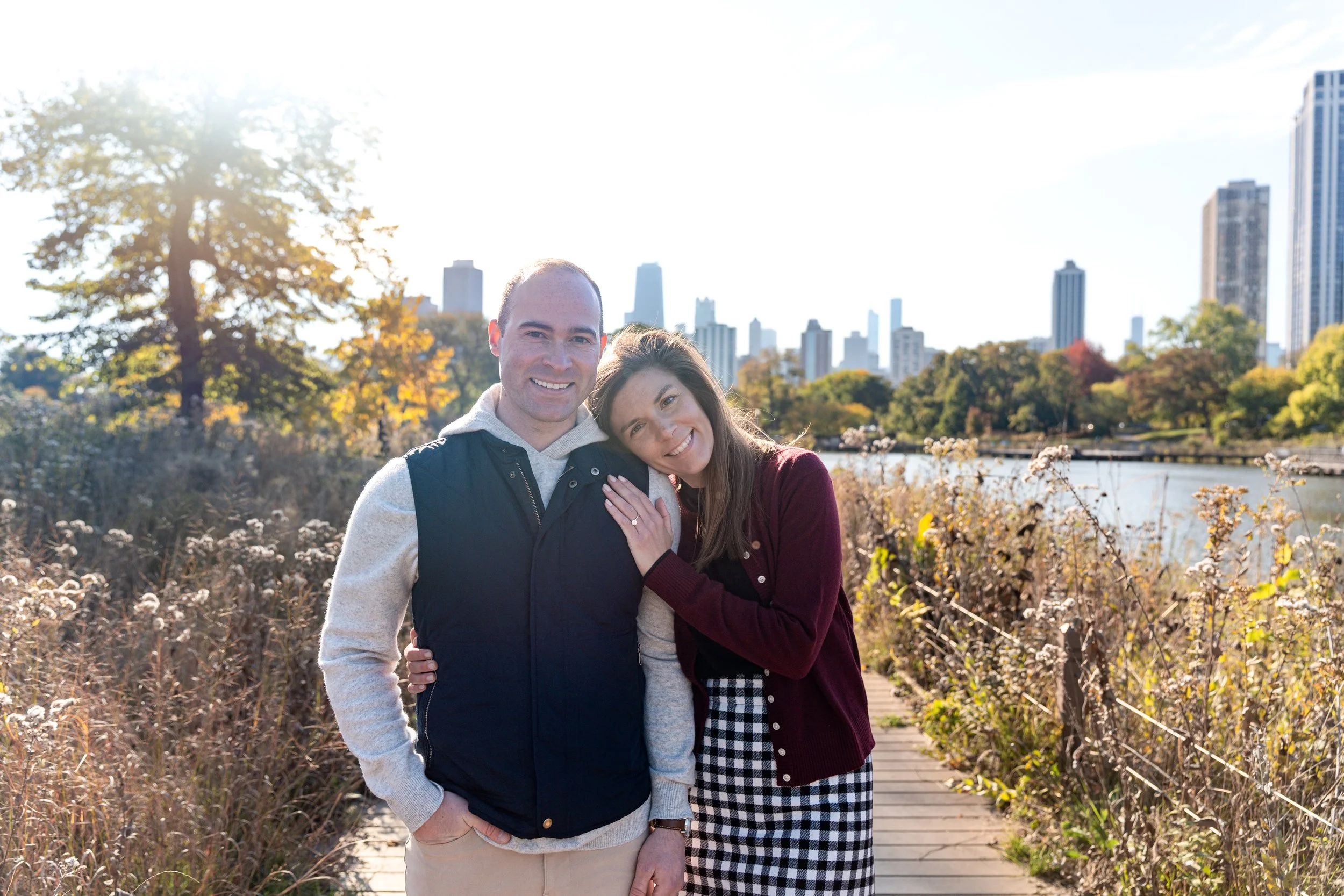 A smiling man and woman standing close together on a park pathway beside a river in an urban setting with tall buildings and trees in the background on a sunny day.