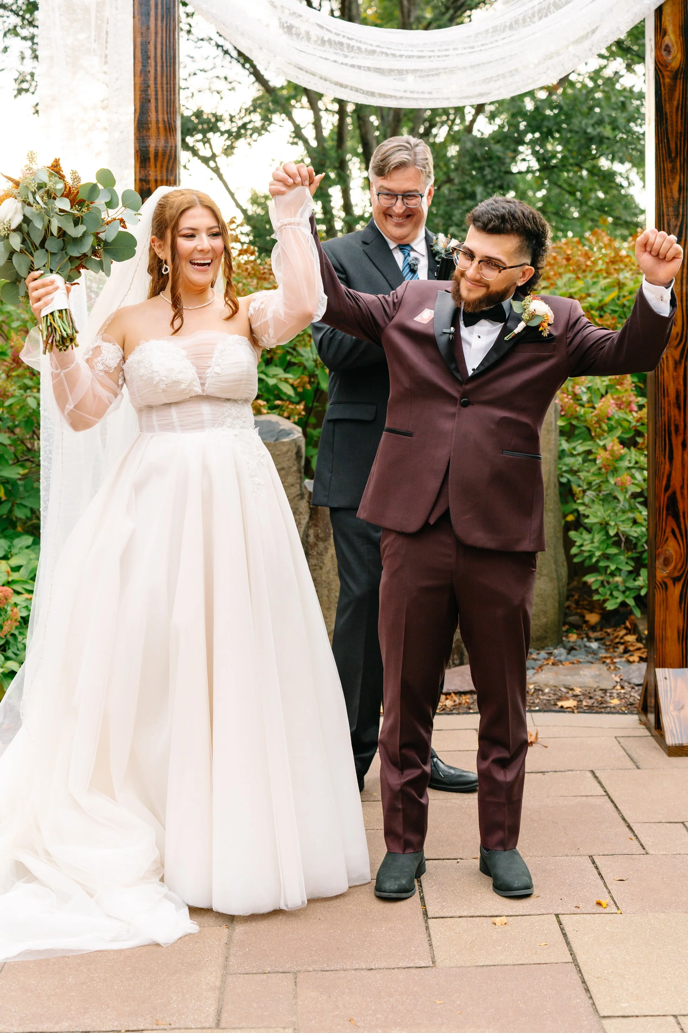A newly married couple celebrating their wedding outdoors, with the bride in a white wedding dress holding a bouquet, and the groom in a burgundy tuxedo with a black bow tie, both smiling and raising their fists in victory, accompanied by an offician