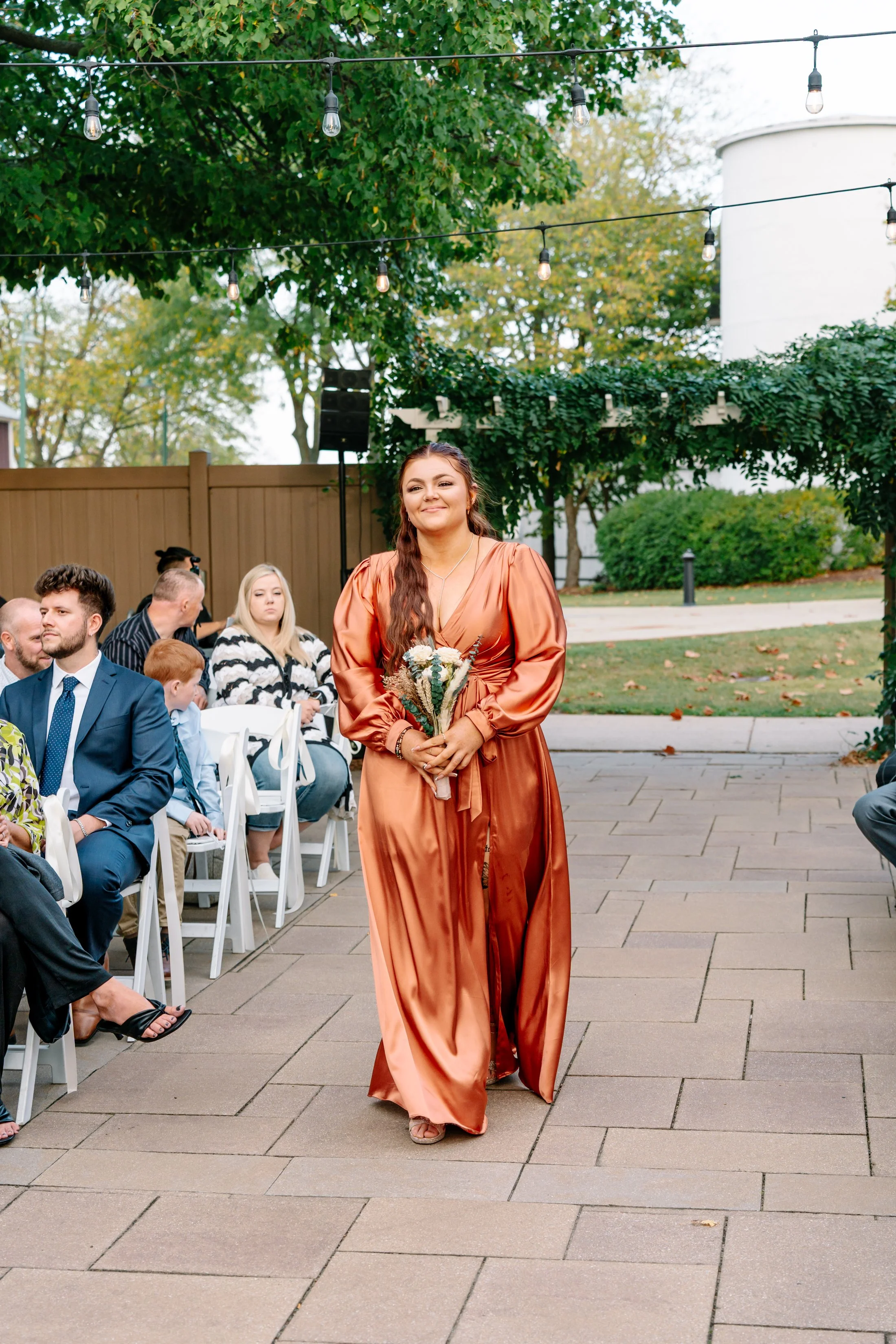 A young woman in a shiny orange dress walking down an outdoor aisle at a wedding, holding a bouquet of flowers, with guests seated on either side.