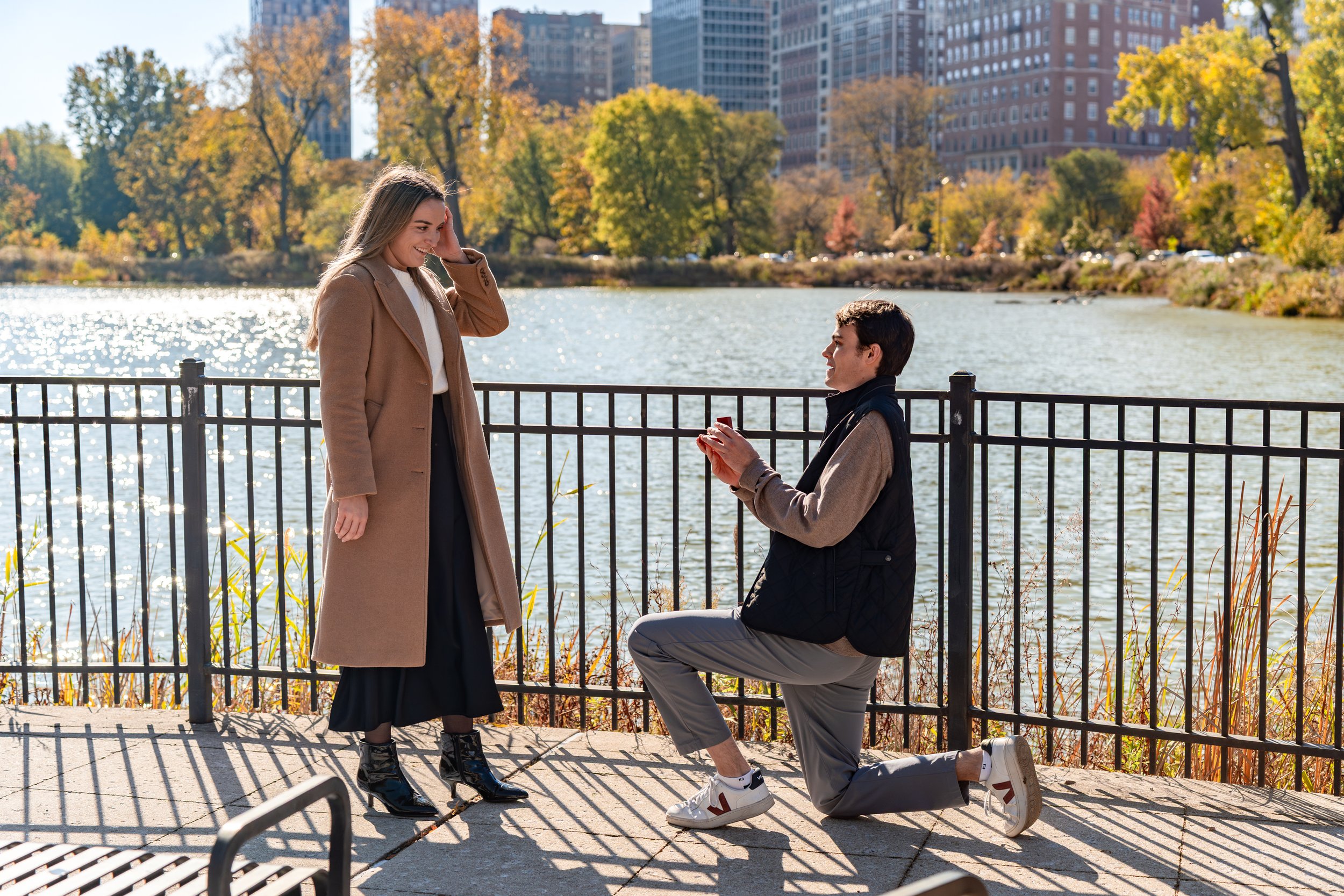 A man on one knee proposing to a woman by a lake in a city park during fall, with colorful trees and tall buildings in the background.