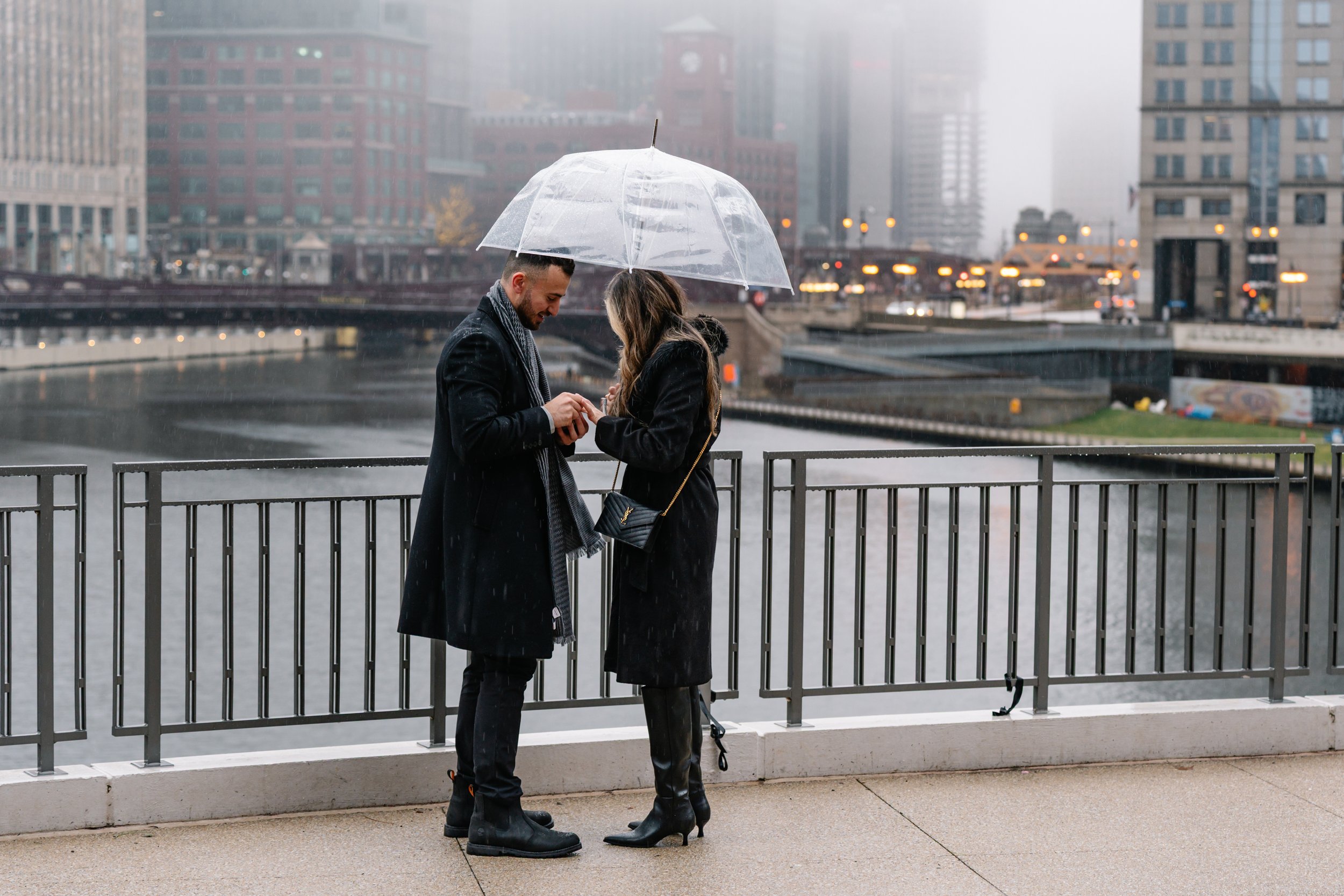 A man and woman standing on a city sidewalk under a transparent umbrella in the rain, looking at a phone together, with a river and city buildings in the background.