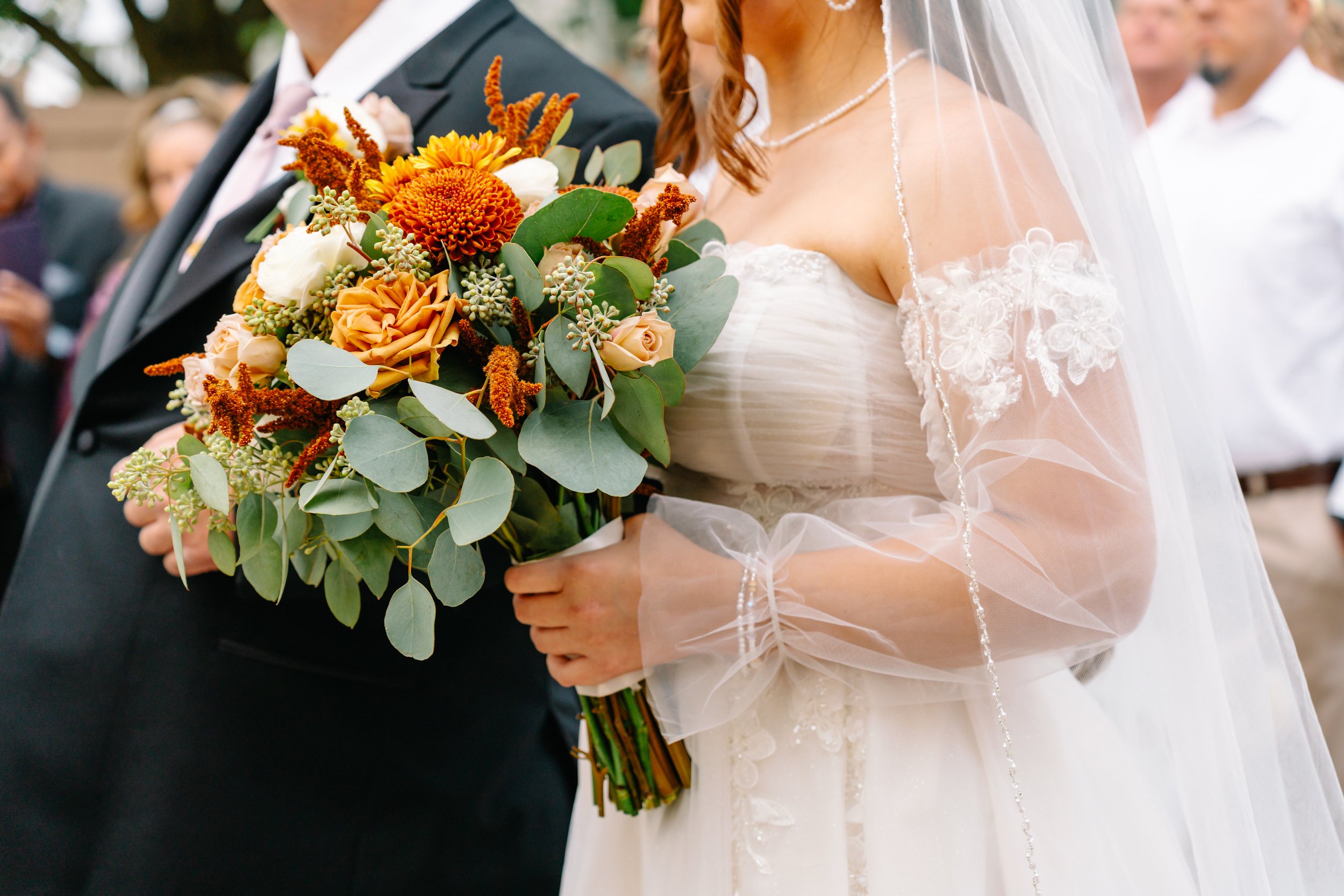 Close-up of a bride holding a bouquet with orange, white, and peach flowers, and an older man in a black suit standing nearby during a wedding ceremony.