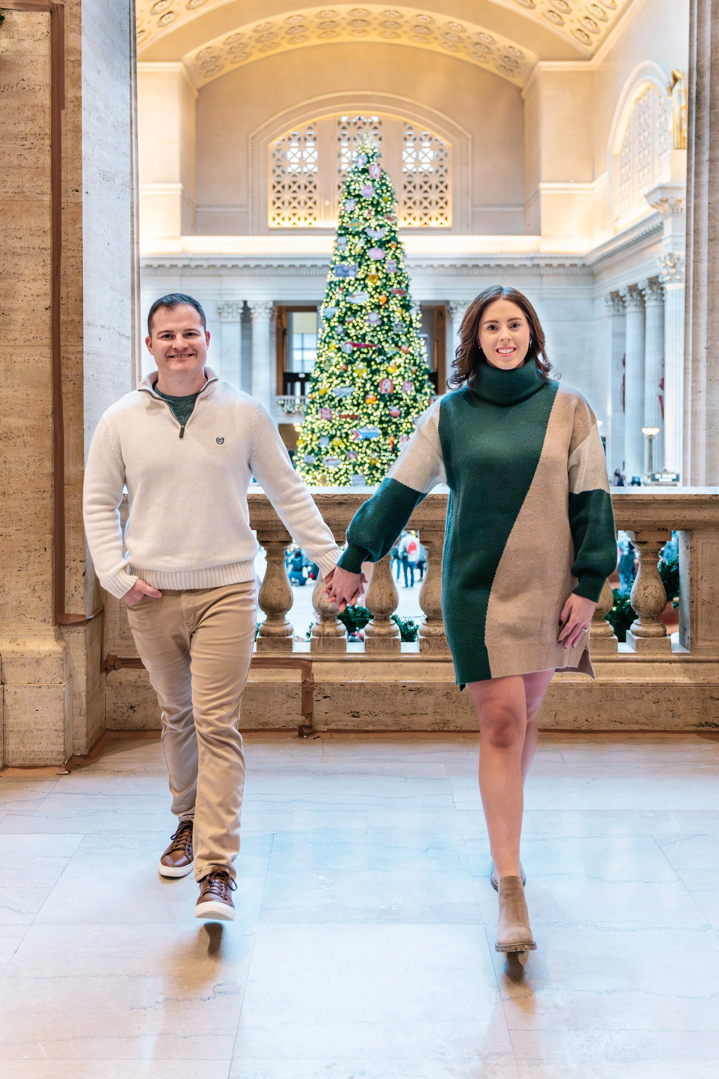 A man and woman holding hands inside a grand hall with a Christmas tree in the background.