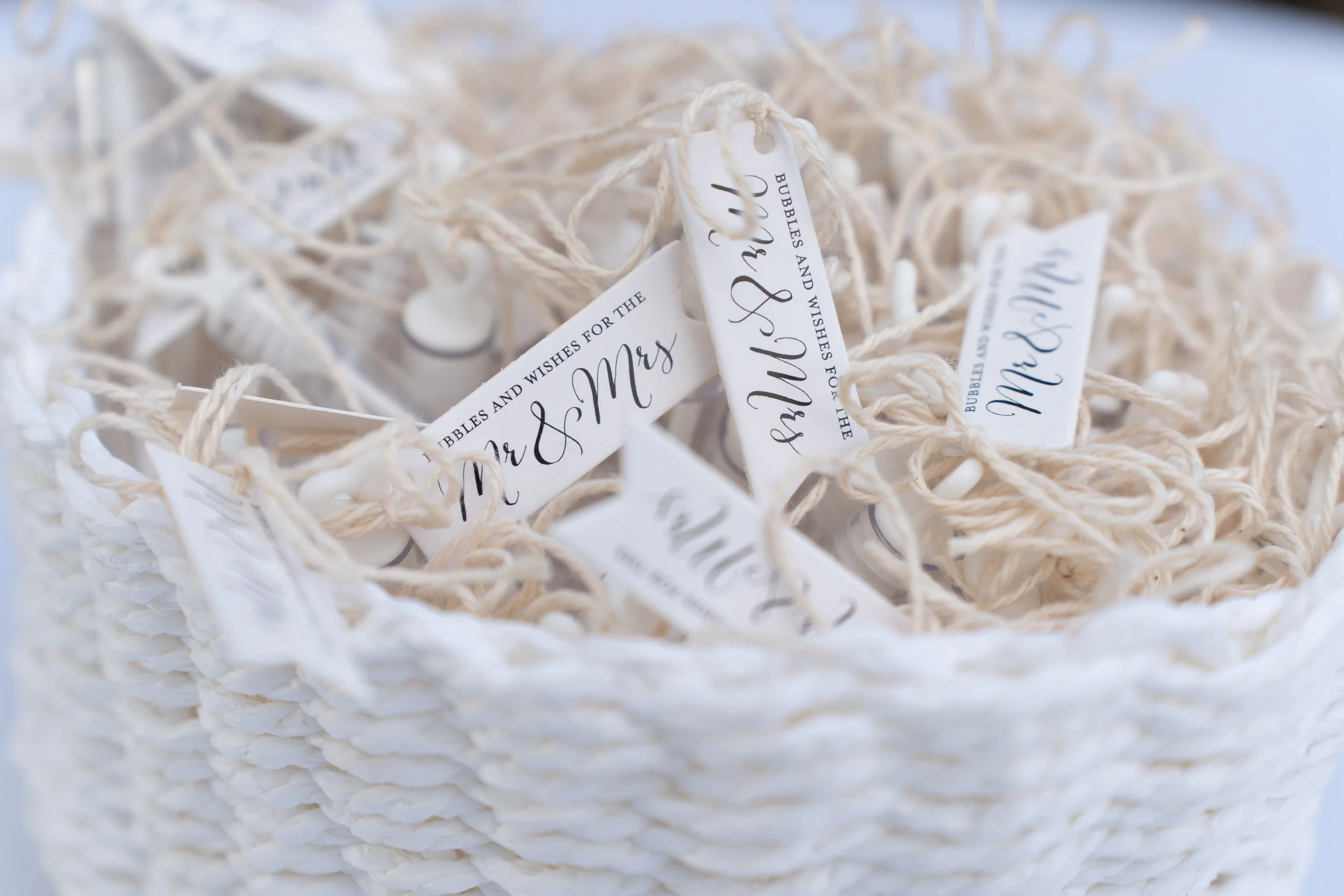 A white basket filled with small get-well-soon gifts tied with beige string and white tags with black cursive text that says 'Wishes & Bubbles for the Mr & Mrs'.