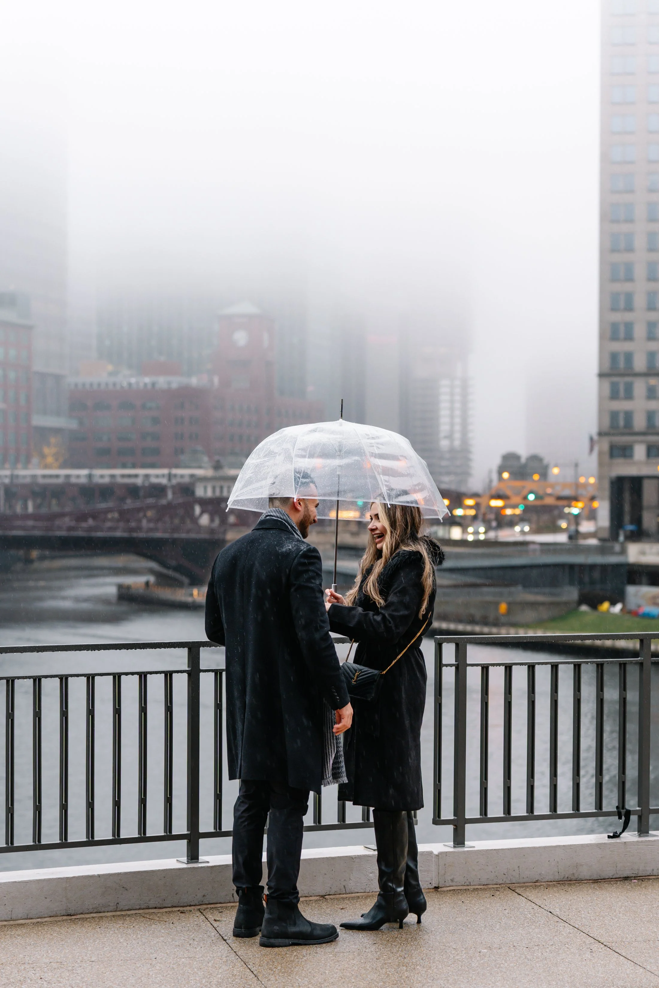 A couple under a clear umbrella sharing a laugh on a rainy city bridge, with foggy skyscrapers and a river in the background.