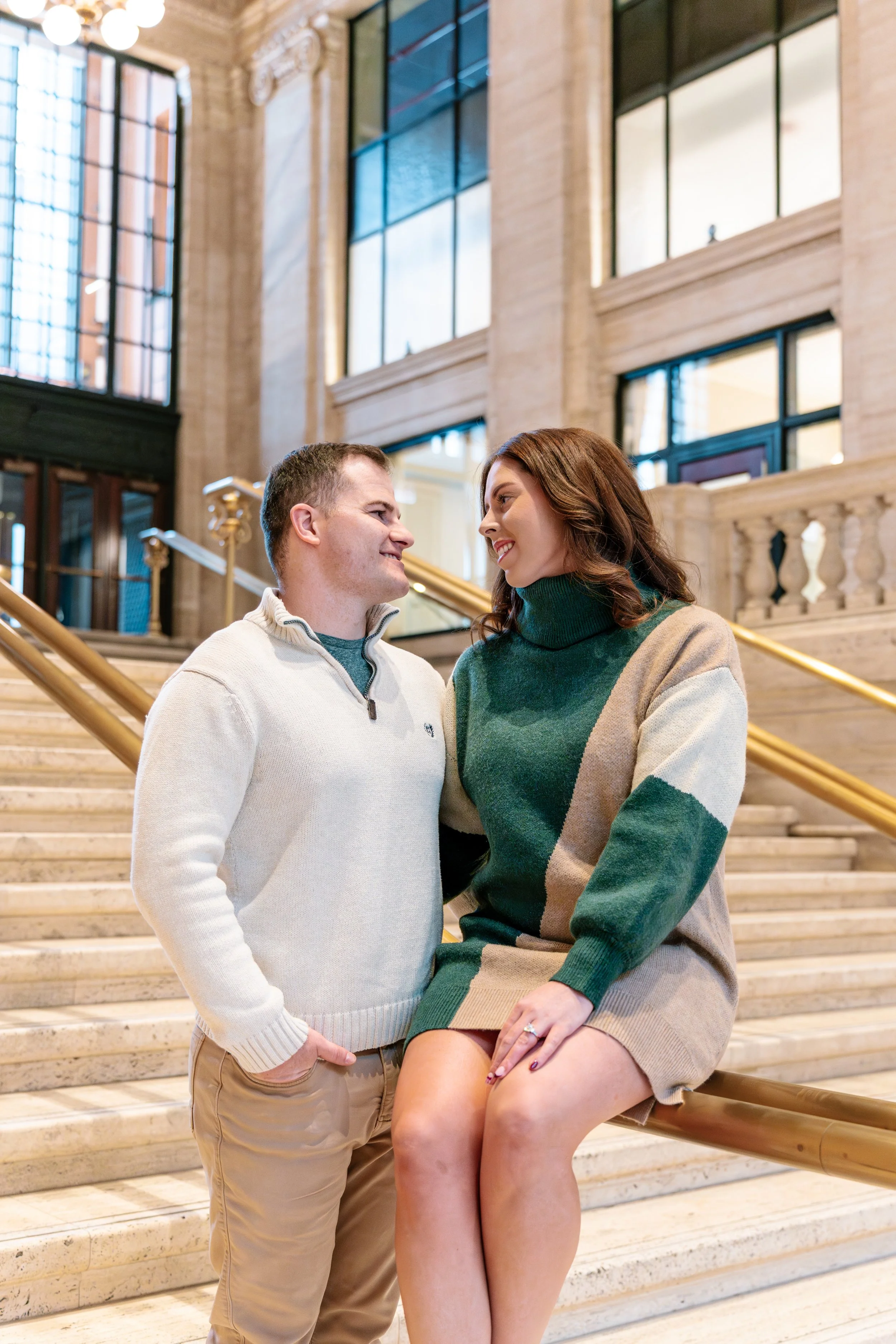 A man and woman smiling at each other on a staircase inside a grand, well-lit building with large windows and stone architecture.