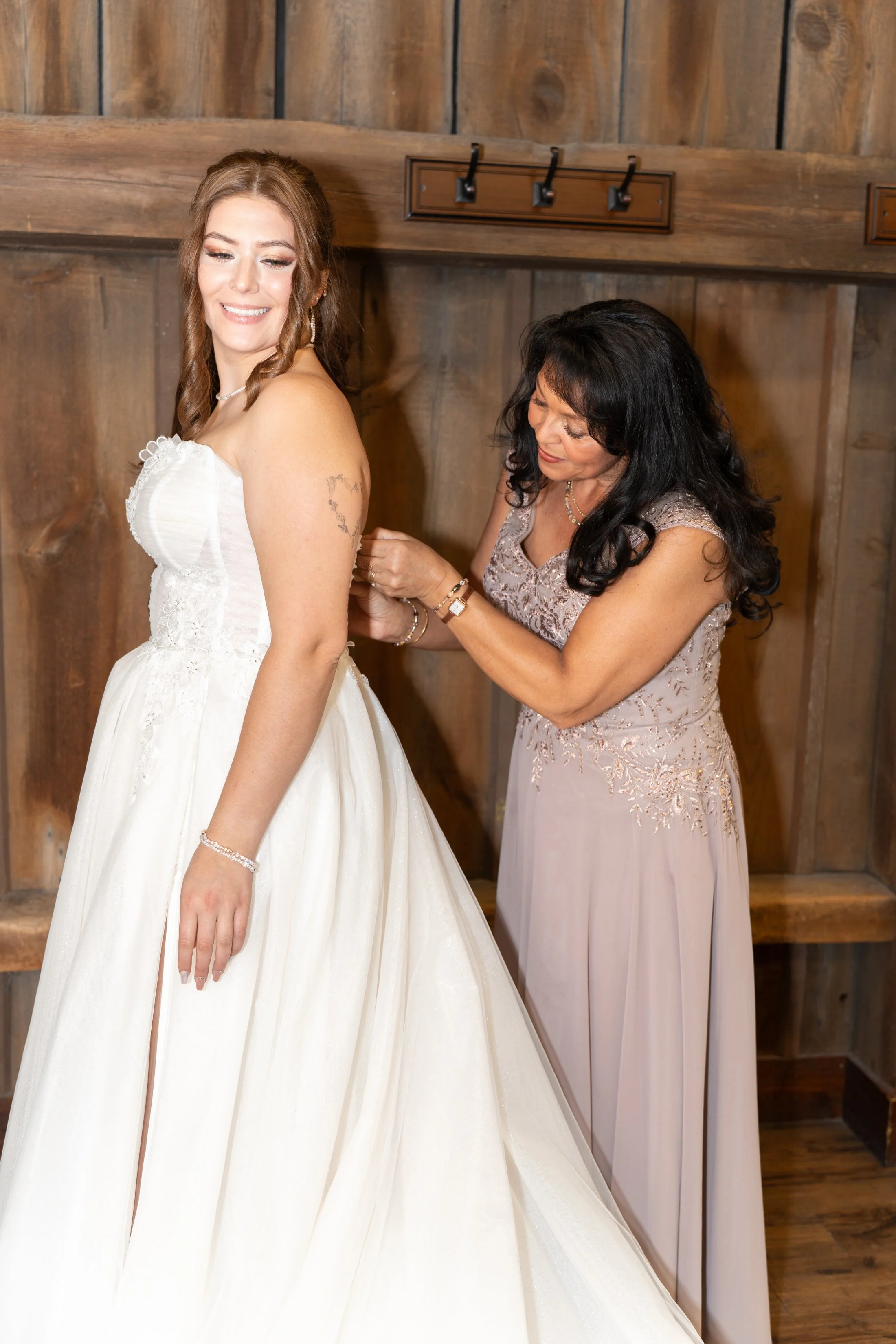 A bride in a white wedding dress is being helped with her dress by a woman in a lavender gown, standing in front of a wooden wall.