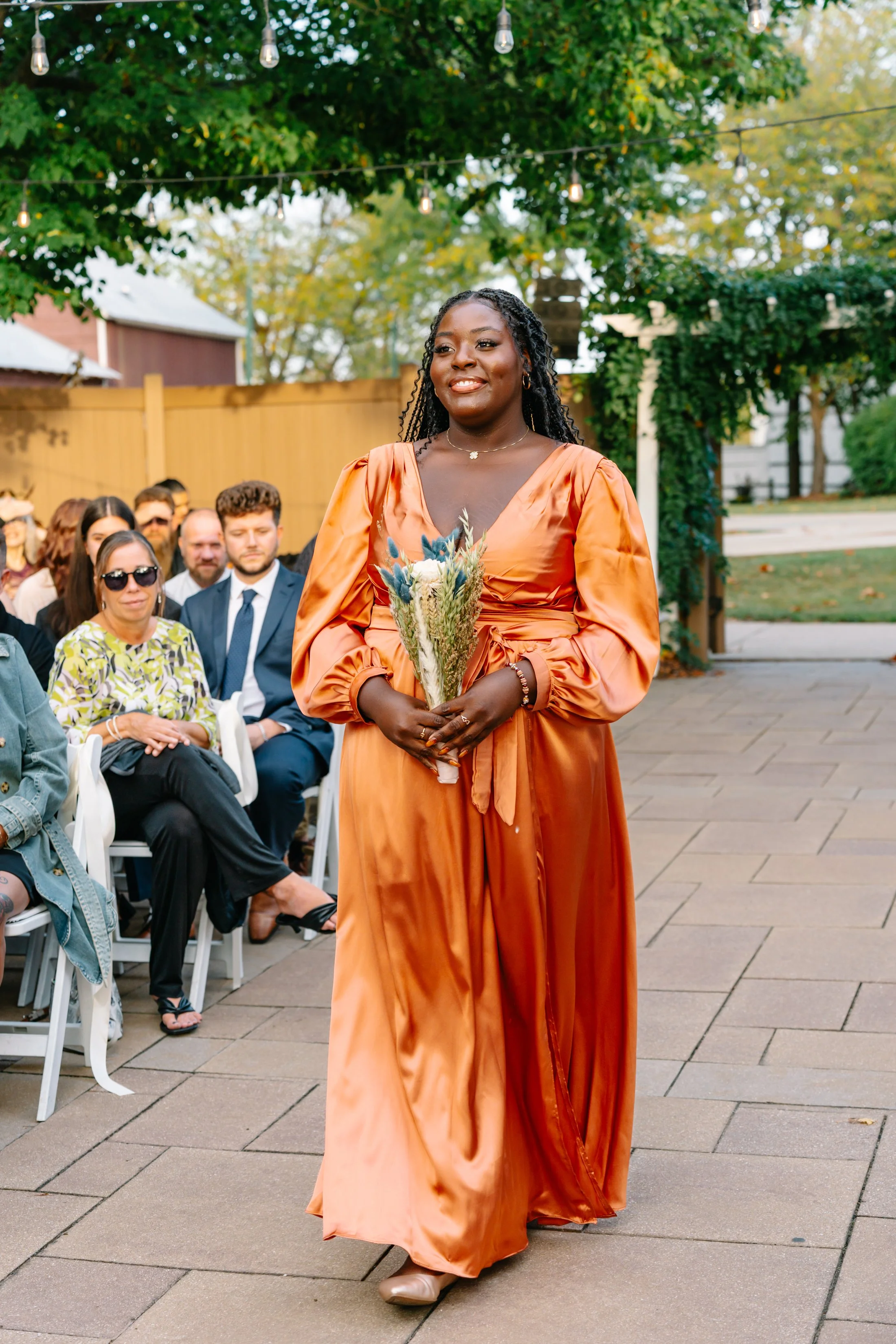 A woman in an orange satin dress holding a bouquet of flowers at an outdoor event, with seated guests in the background and string lights overhead.