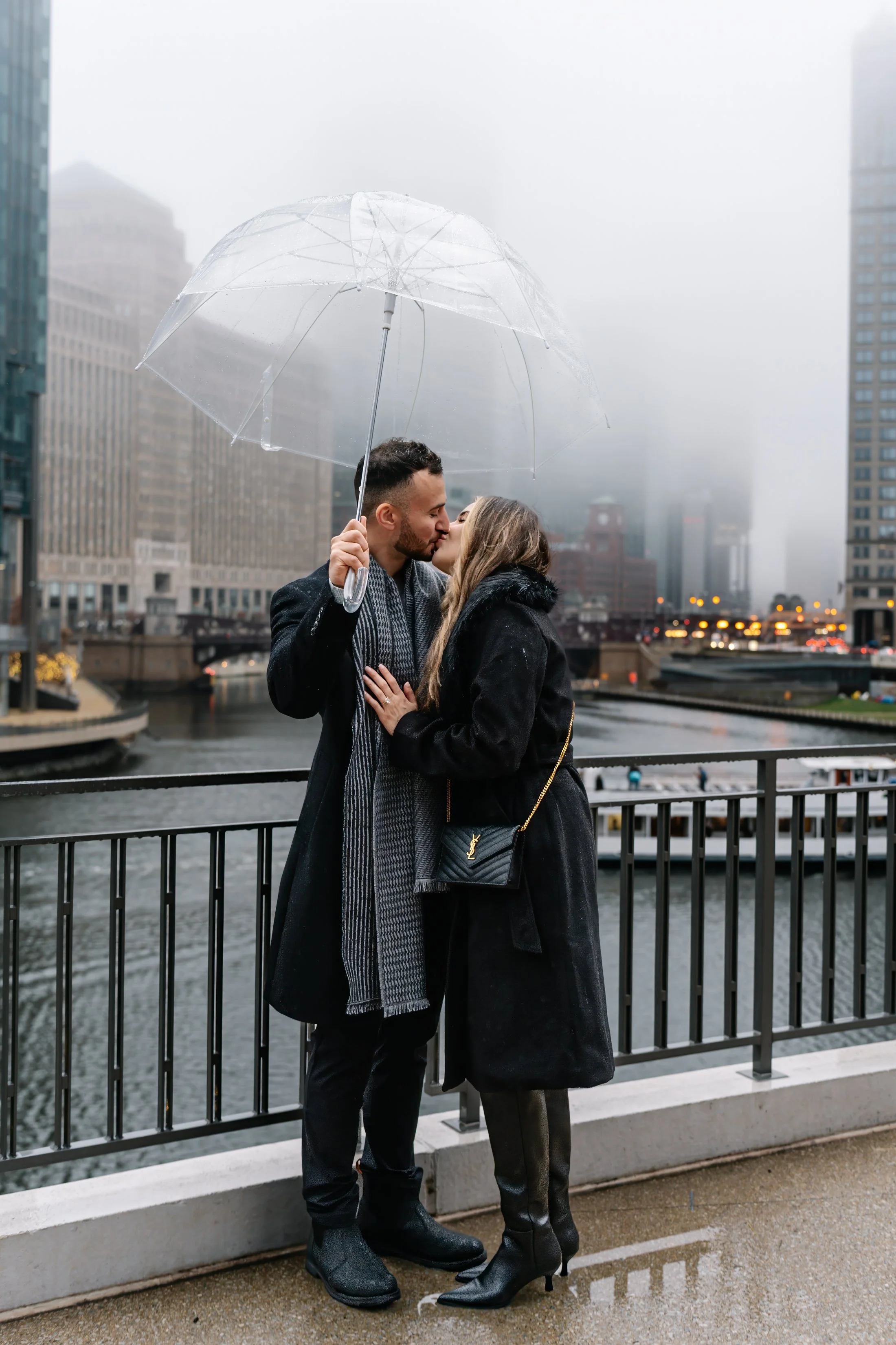 A couple sharing a kiss under an umbrella in a city with tall buildings and a river, on a rainy day.