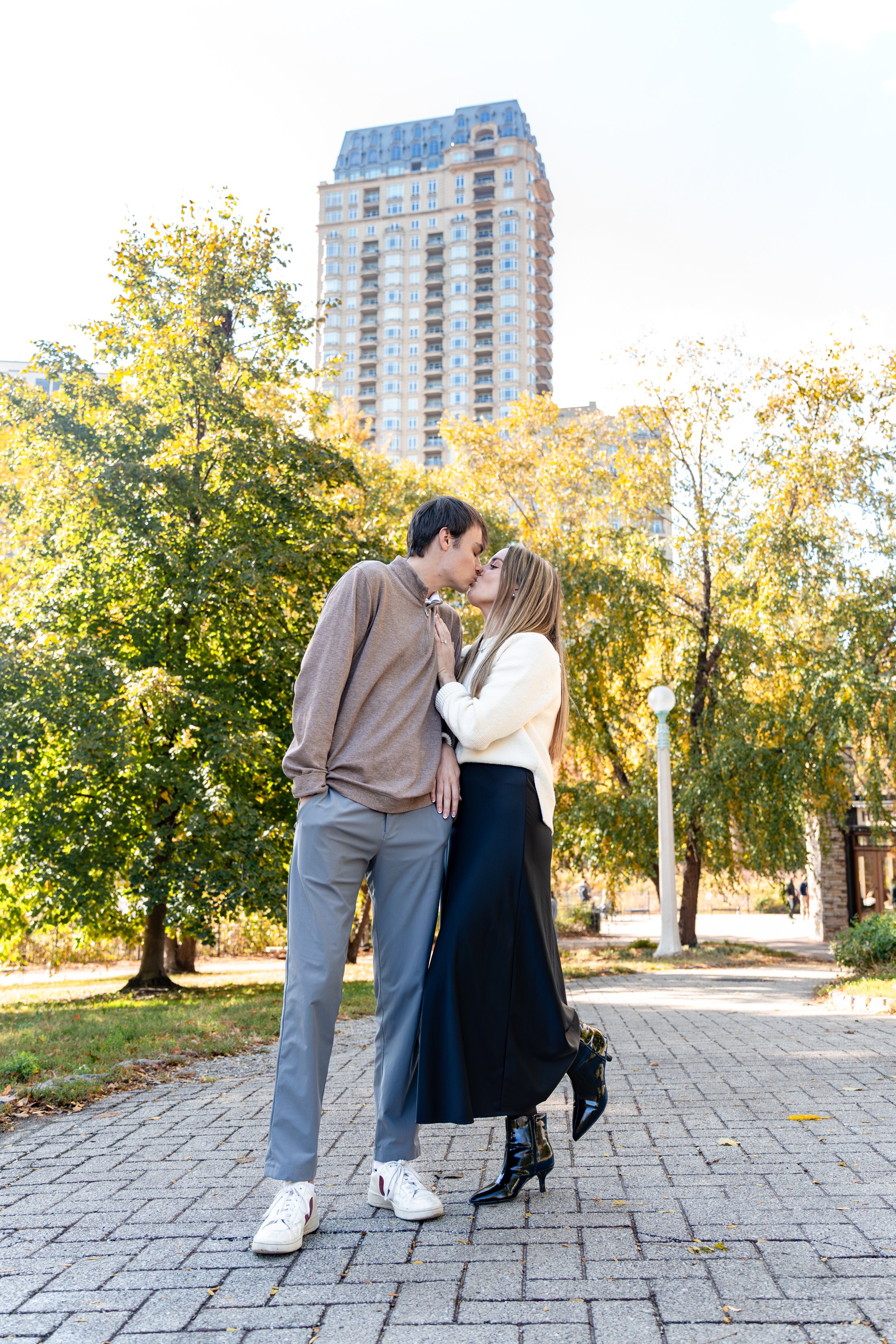 A couple sharing a kiss outdoors in a park during daytime, with trees and a tall building in the background.