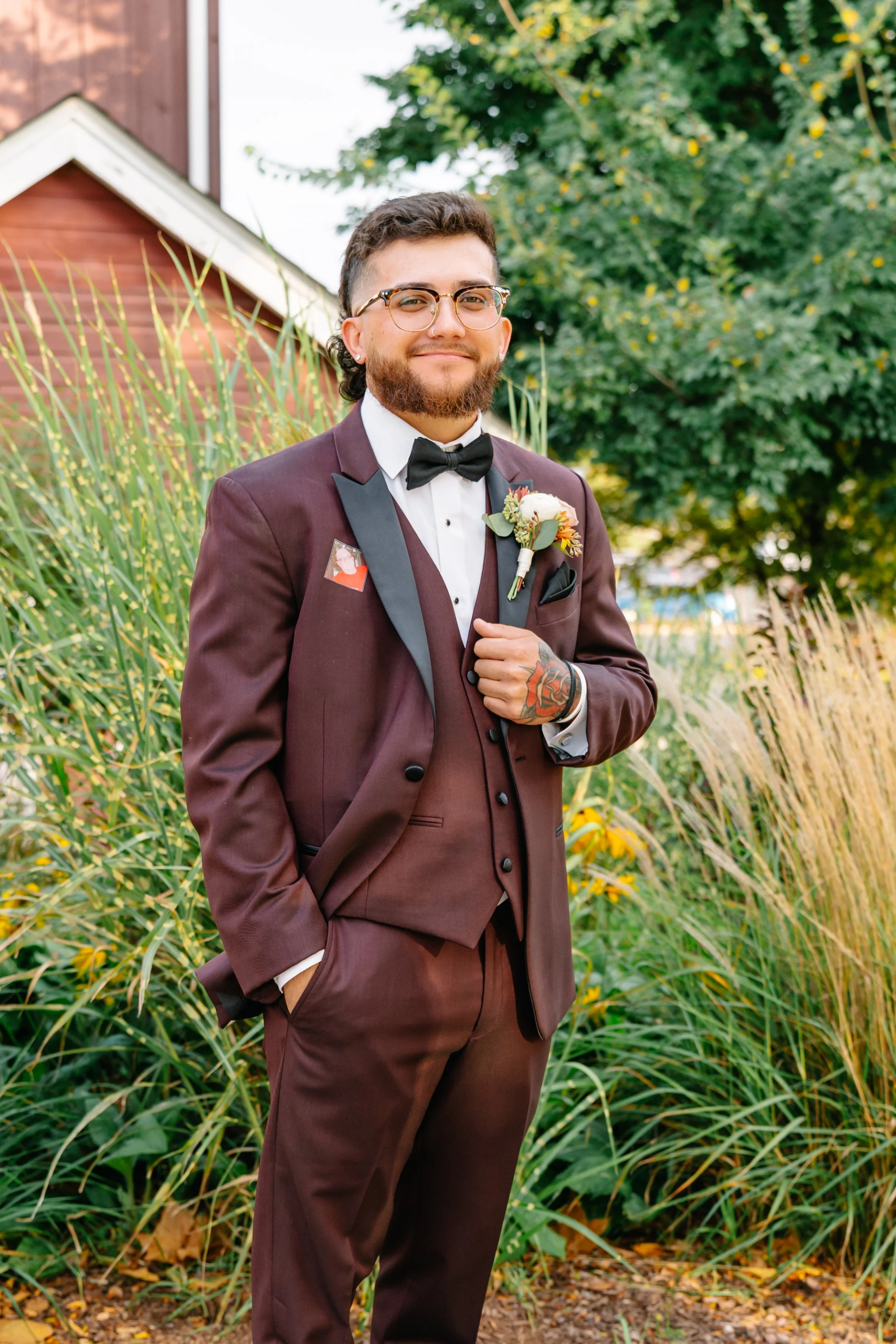 Man in a brown suit with black lapel, white shirt, black bow tie, and glasses, standing outdoors near green plants and trees, with a small boutonniere on his lapel.
