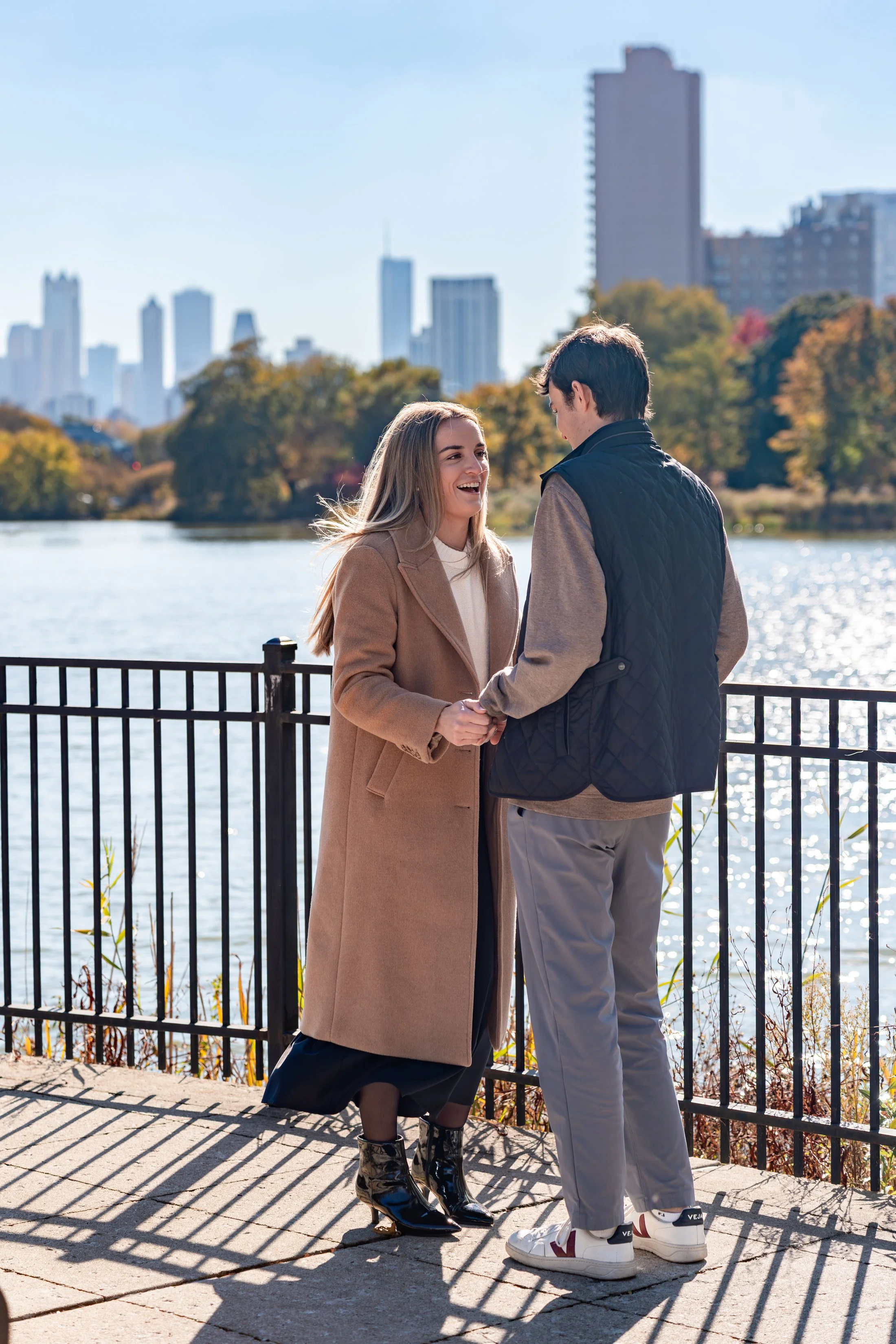 A young couple holding hands and smiling by a river with a city skyline in the background on a sunny day.