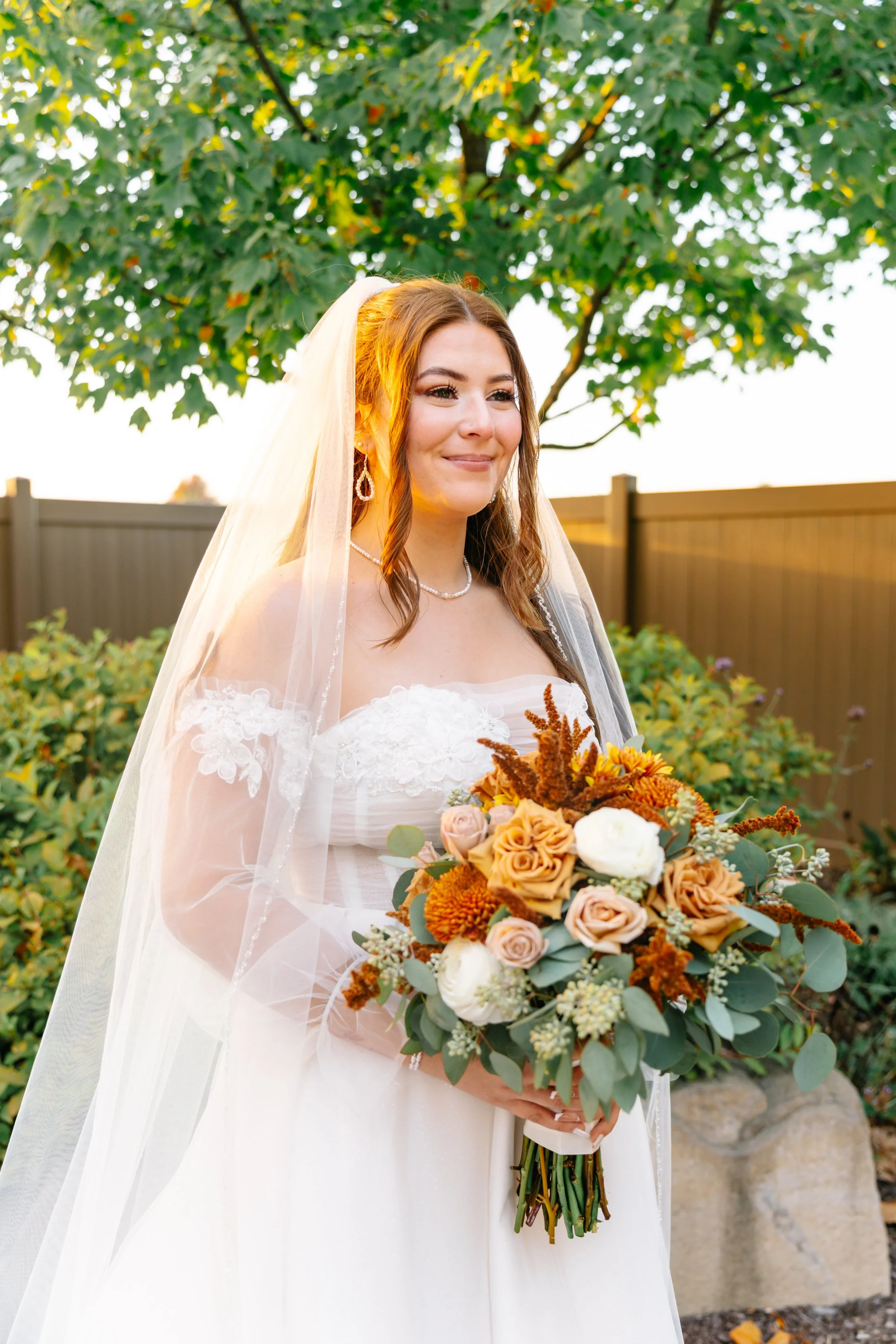 A bride holding a bouquet of flowers on her wedding day, standing outdoors in front of a green leafy tree and a wooden fence during sunset.