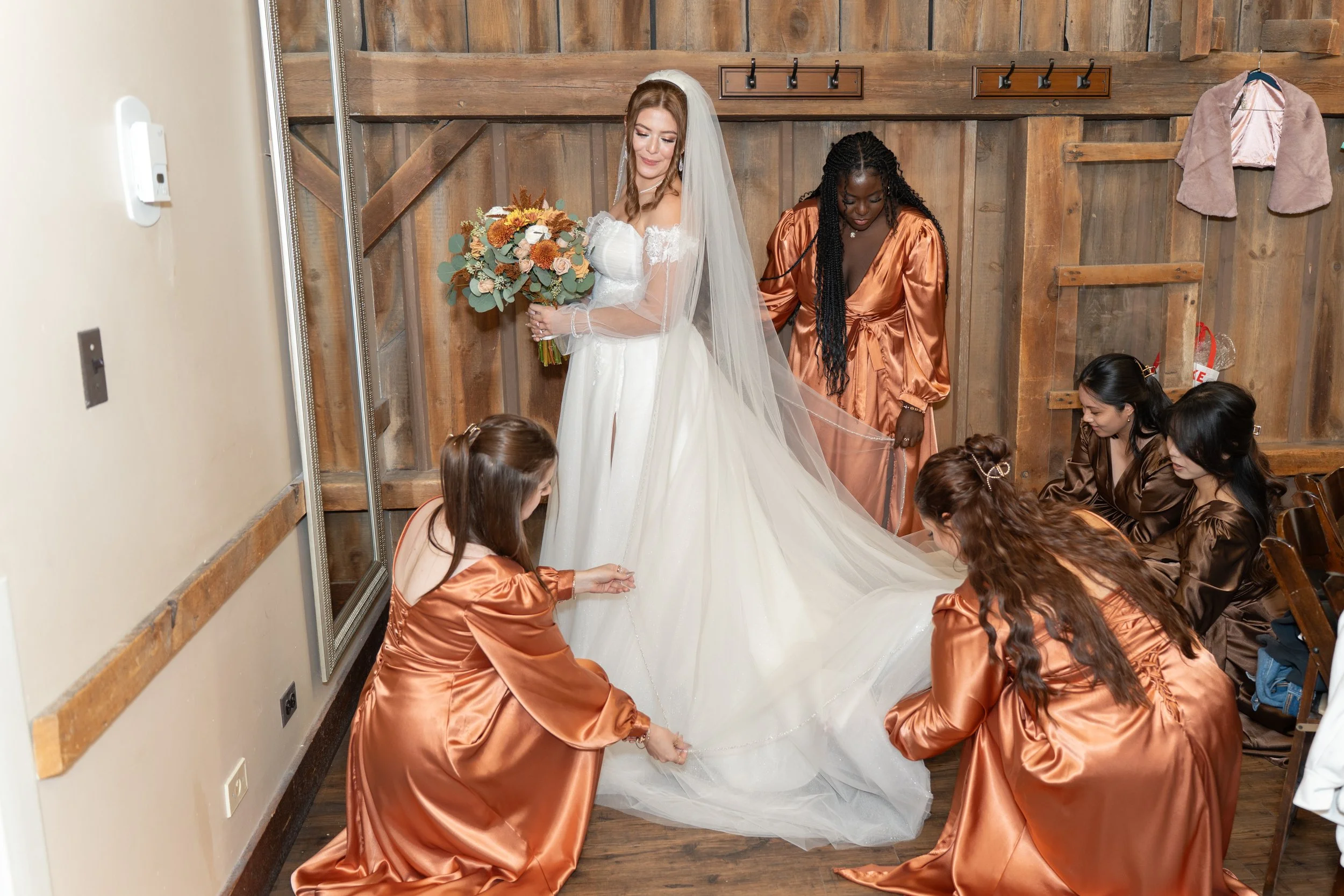 A bride in a white wedding dress with a veil holding a bouquet of flowers, standing as her bridesmaids in bronze satin robes helps with her train in a rustic wooden room with a small wall-mounted coat rack.