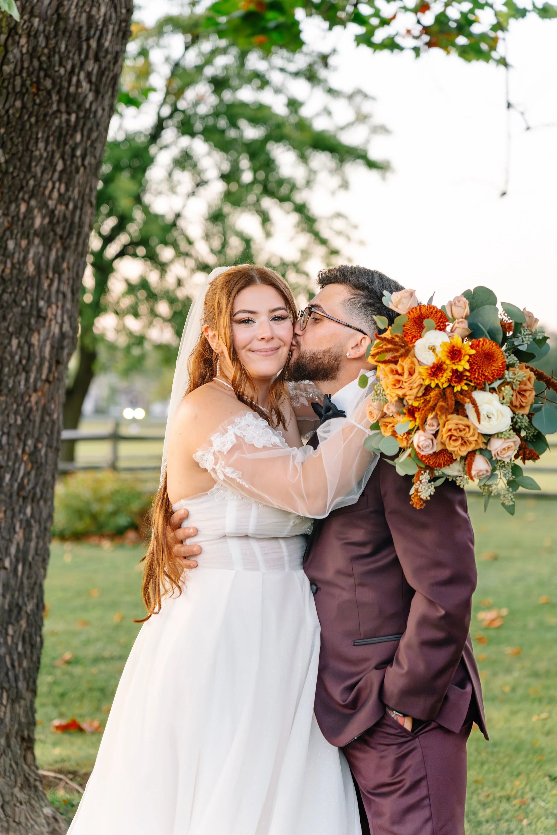 A bride and groom embracing outdoors during their wedding, the groom kissing the bride's cheek and holding a large bouquet of orange and white flowers, with green leaves, near a tree.