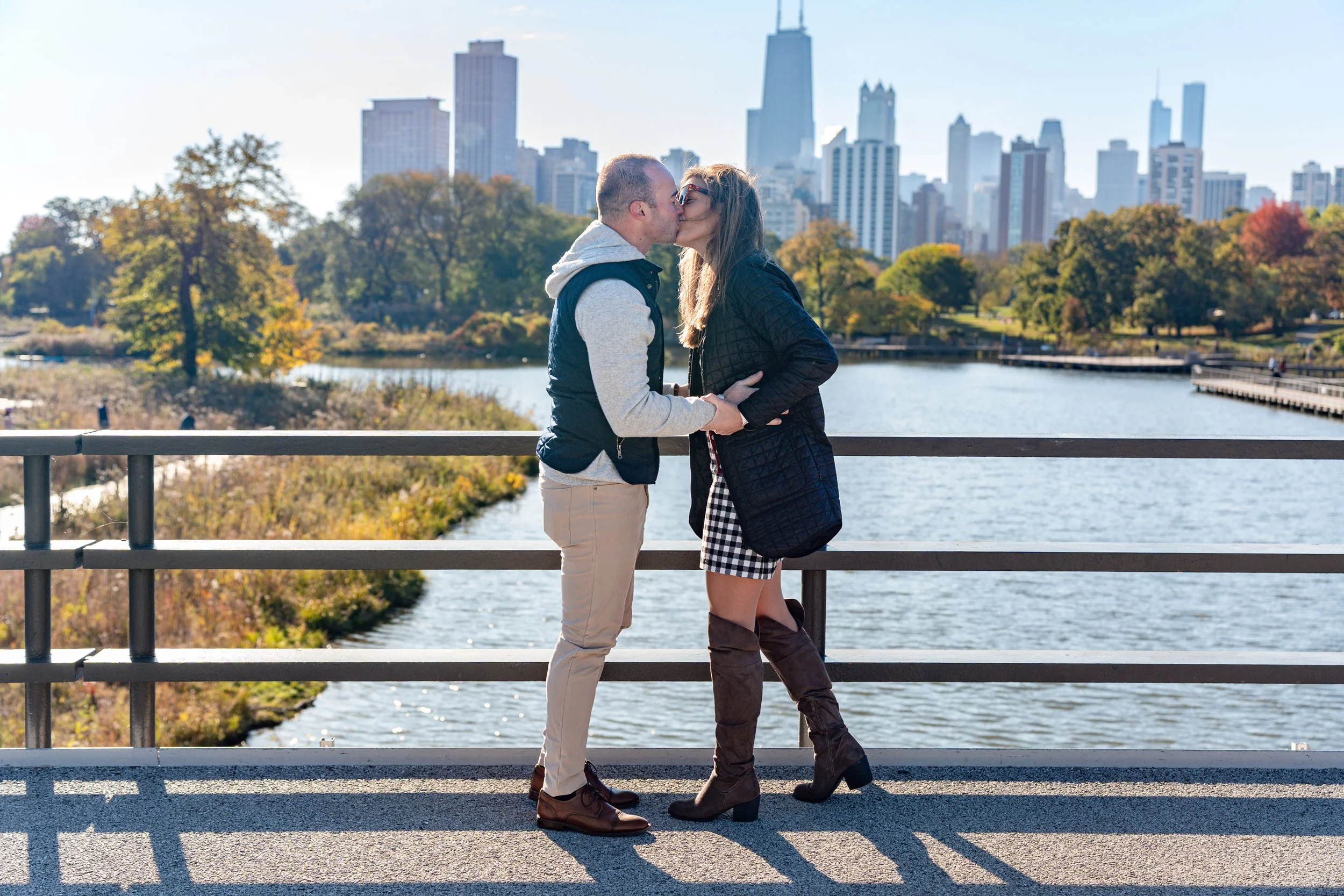 A couple shares a kiss on a bridge with a city skyline and park in the background during fall.