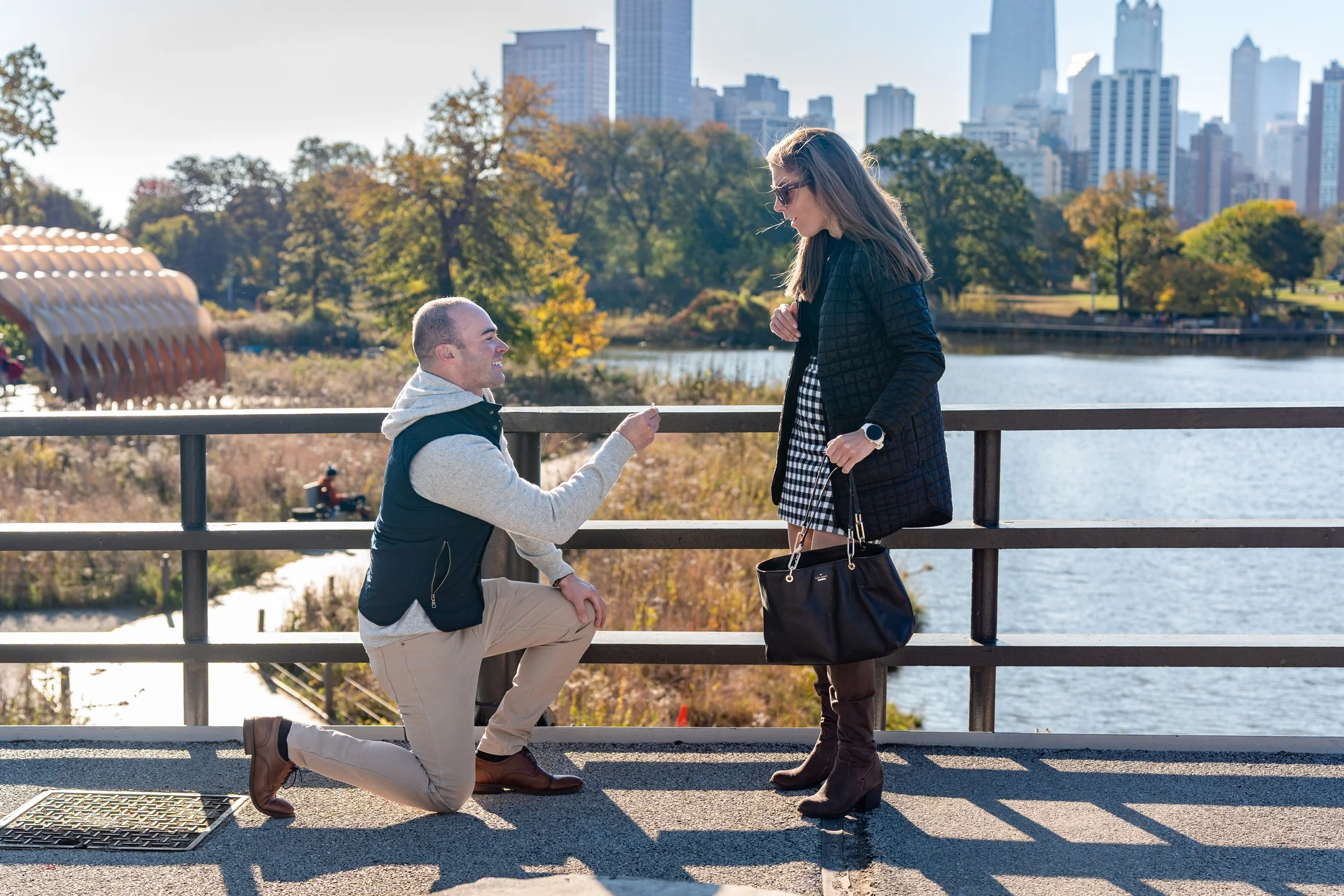 A man proposing marriage to a woman by kneeling on one knee on a bridge near a river with city skyline and trees in the background.