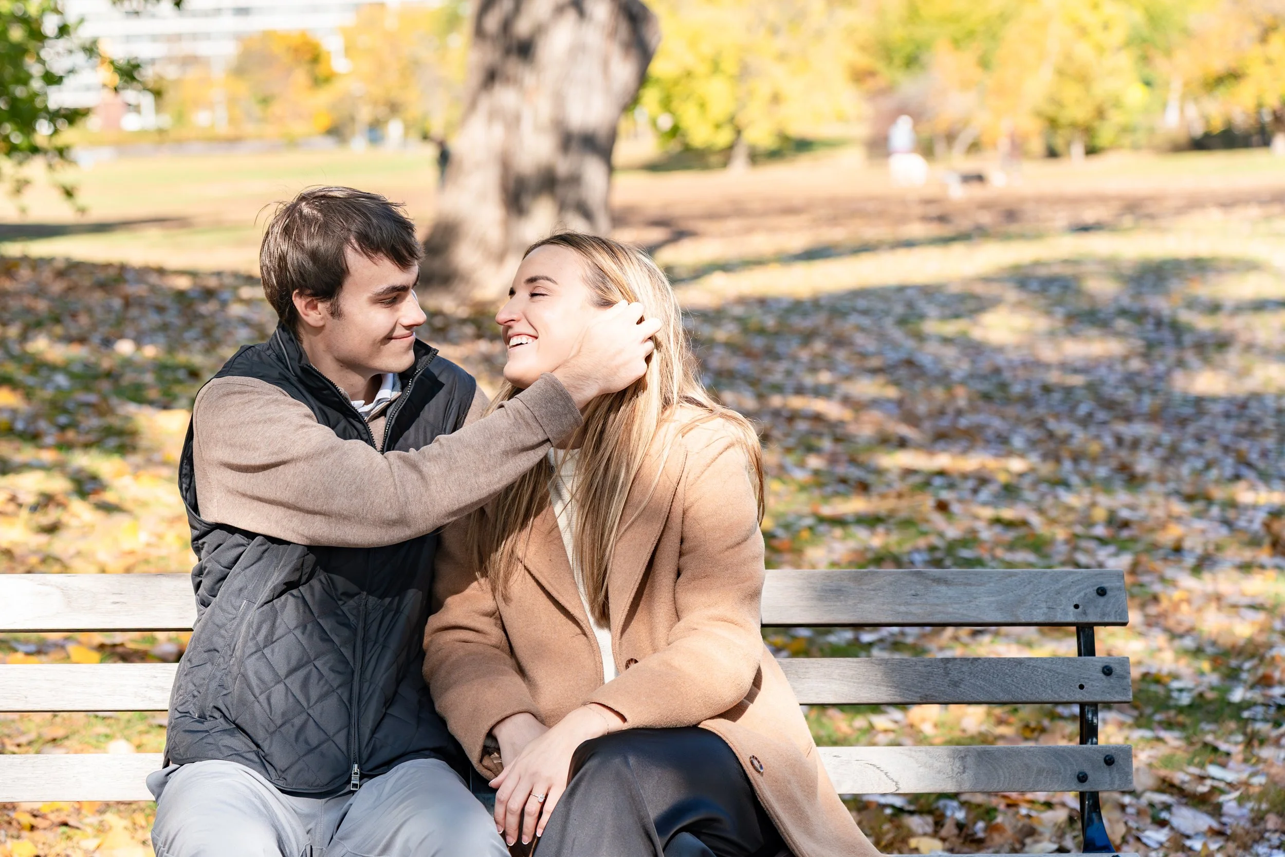 A young man and woman sitting on a park bench, smiling, with fallen leaves and trees in the background.