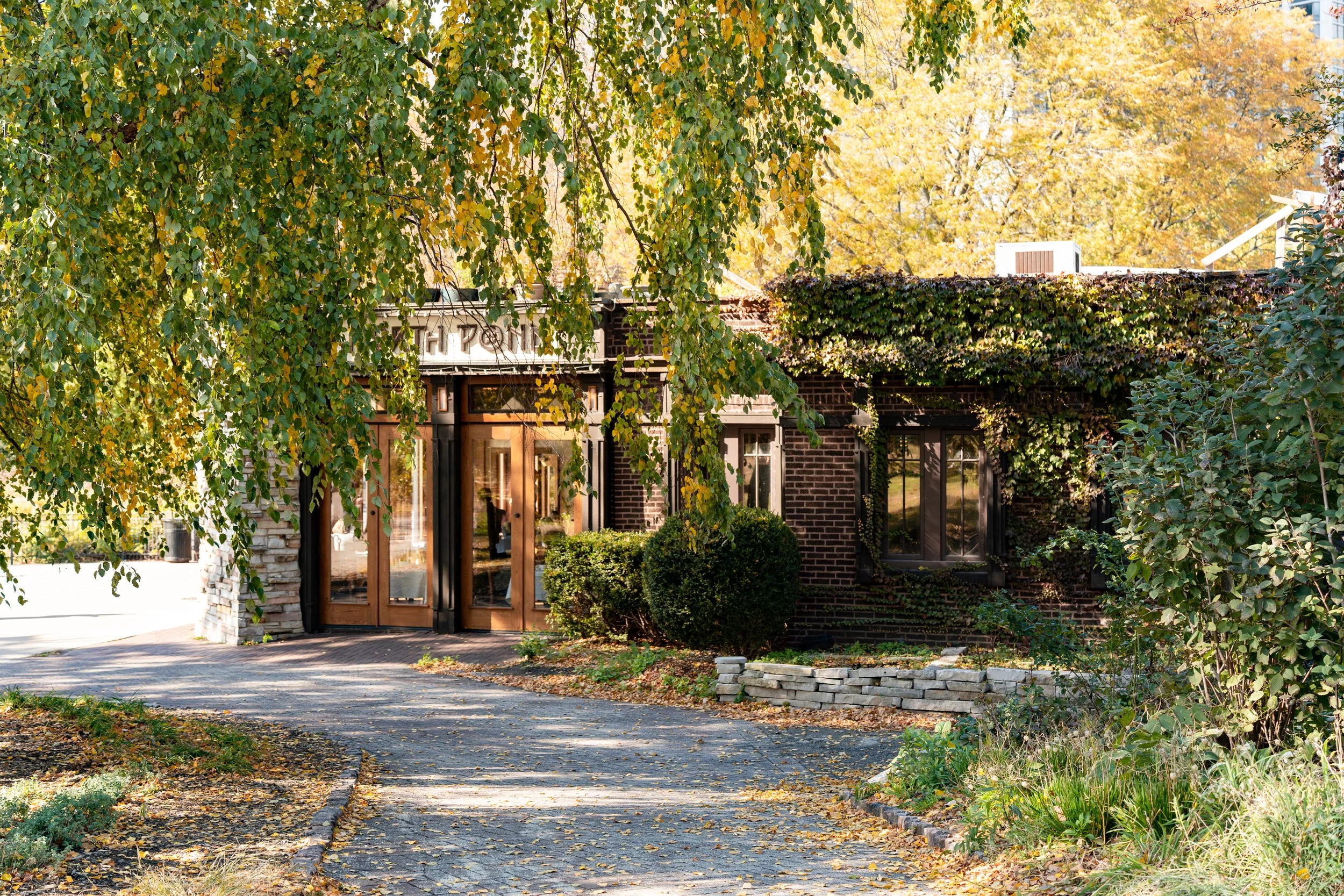 A brick building with large windows, surrounded by lush greenery, trees with fall foliage, and a curved pathway leading to the entrance.