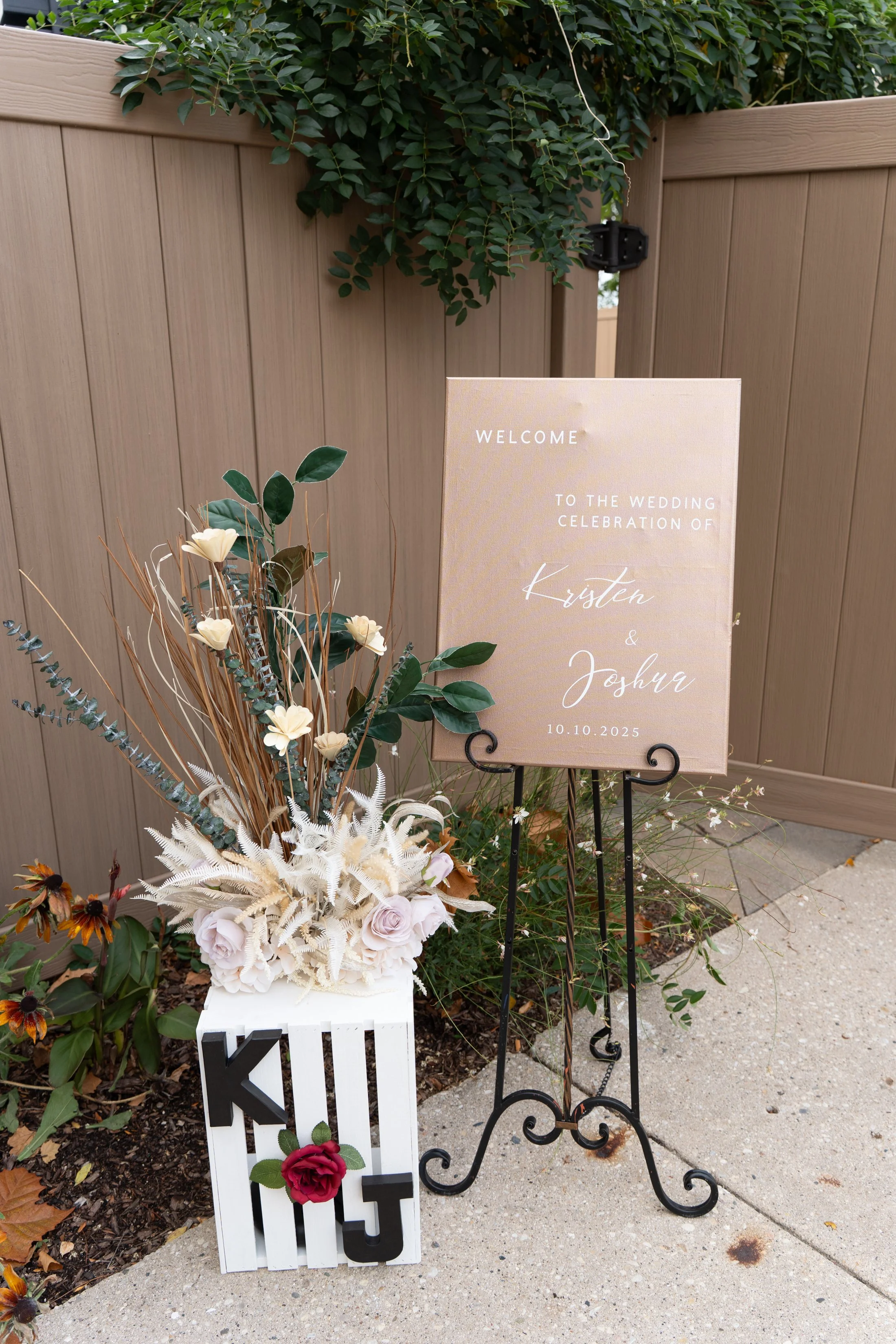 Wedding welcome sign for Kristen and Joshua with date 10.10.2025, placed on a black wrought iron stand next to a floral arrangement with white, blush pink, and green plants, and a white box decorated with the word 'KUT' and a red rose.
