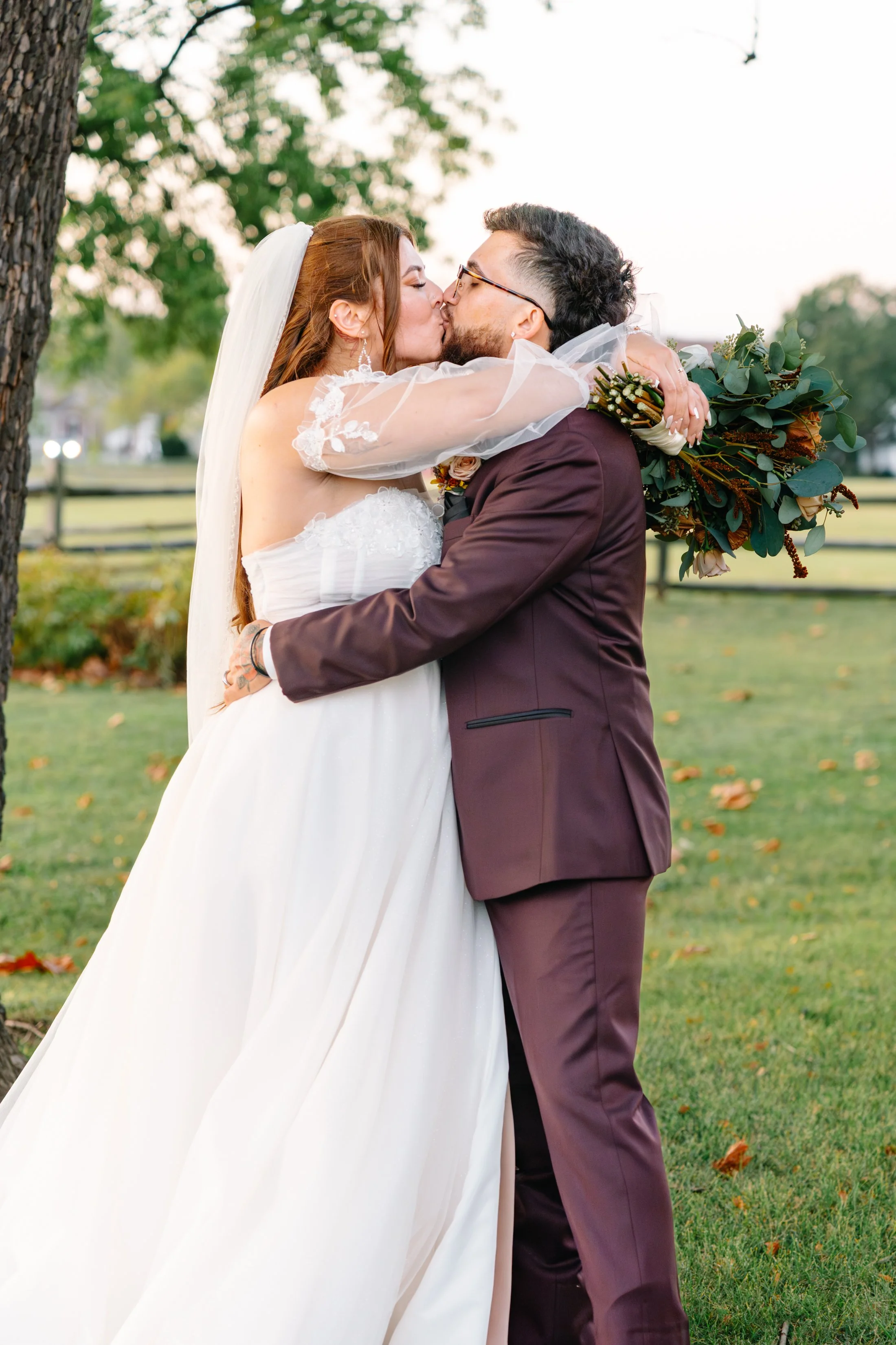A bride and groom kissing outdoors, with the bride in a white wedding dress and veil and the groom in a maroon suit, holding a bouquet of greenery.