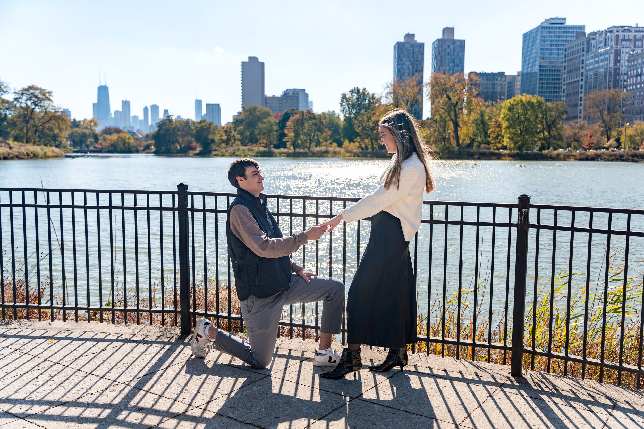A man proposing marriage to a woman by a lake with city skyline in the background during daytime.