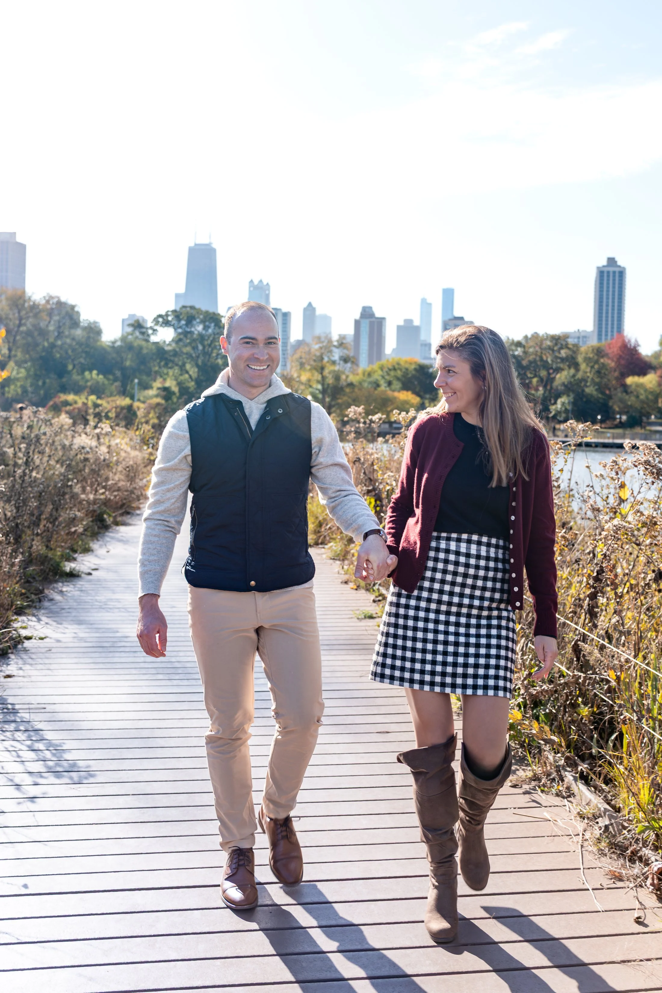 A man and woman holding hands and walking along a wooden pathway in a park with city skyline in the background on a sunny day.