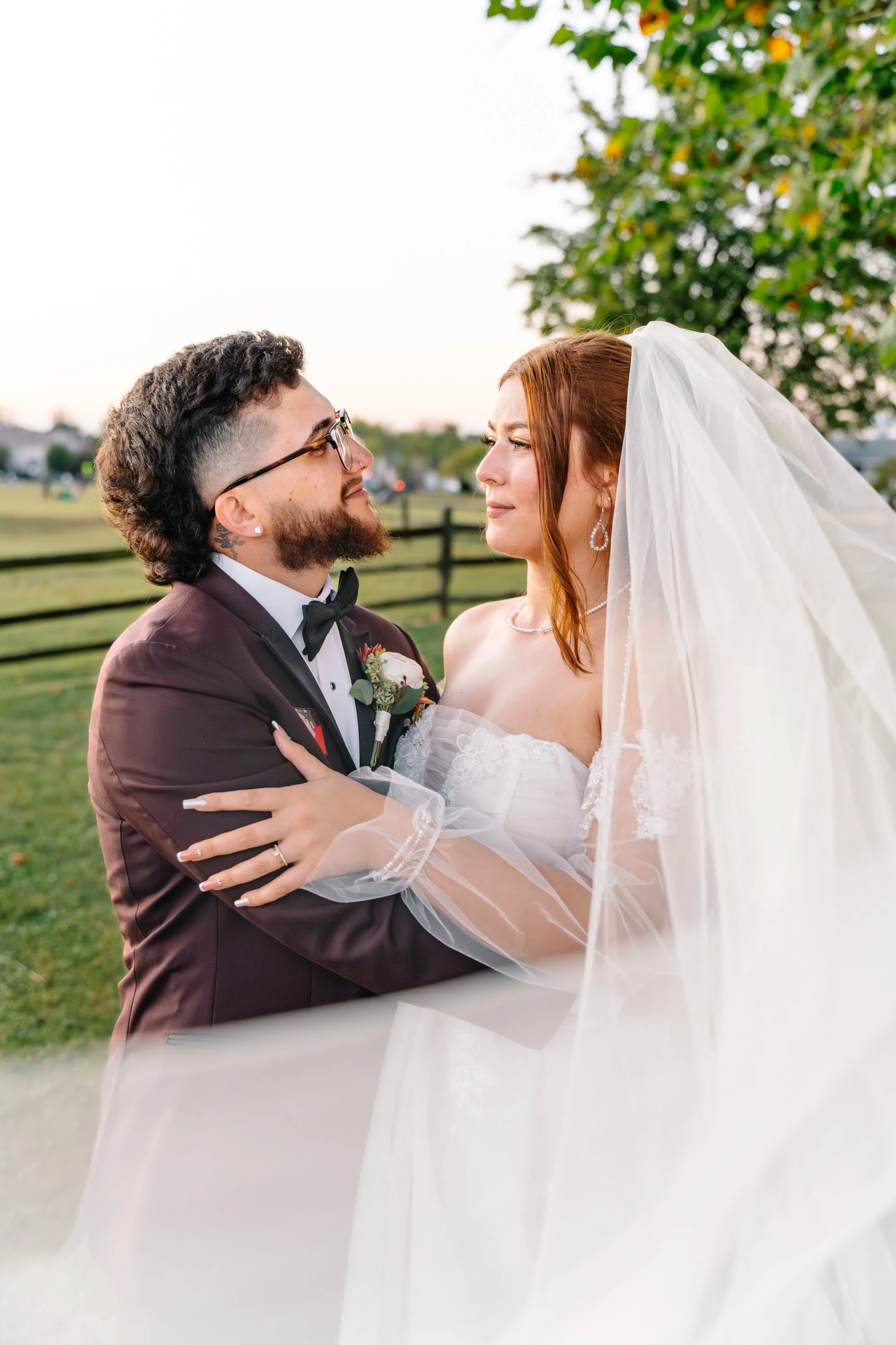 A bride and groom facing each other outdoors at sunset, with a fence and trees in the background. The groom is wearing glasses, a tuxedo, and a boutonniere. The bride is in a white wedding dress with a veil, wearing earrings and a necklace, holding t