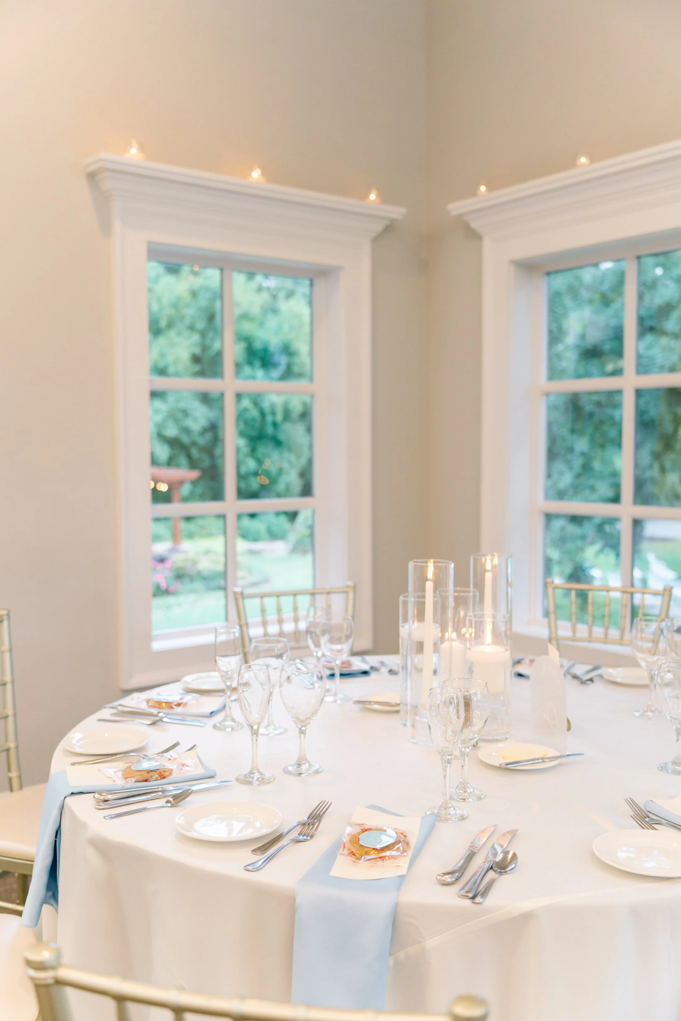 A banquet table set for a formal event with white tablecloth, glassware, plates, cutlery, and candles, in a room with large windows and outside greenery.