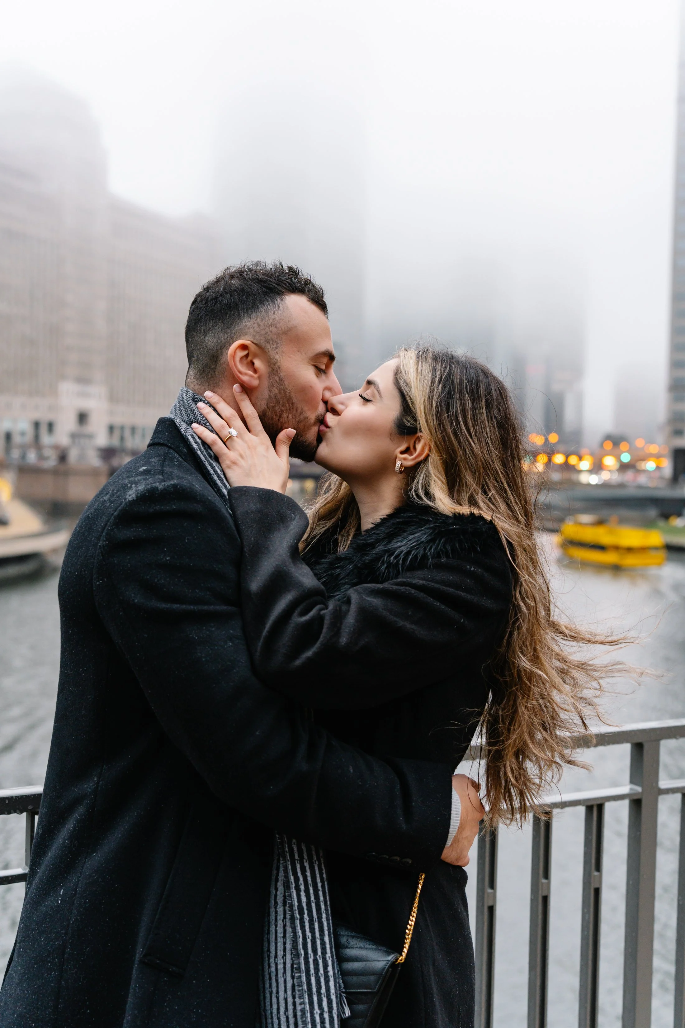 A couple sharing a kiss by a river on a cloudy day, with city buildings in the background.