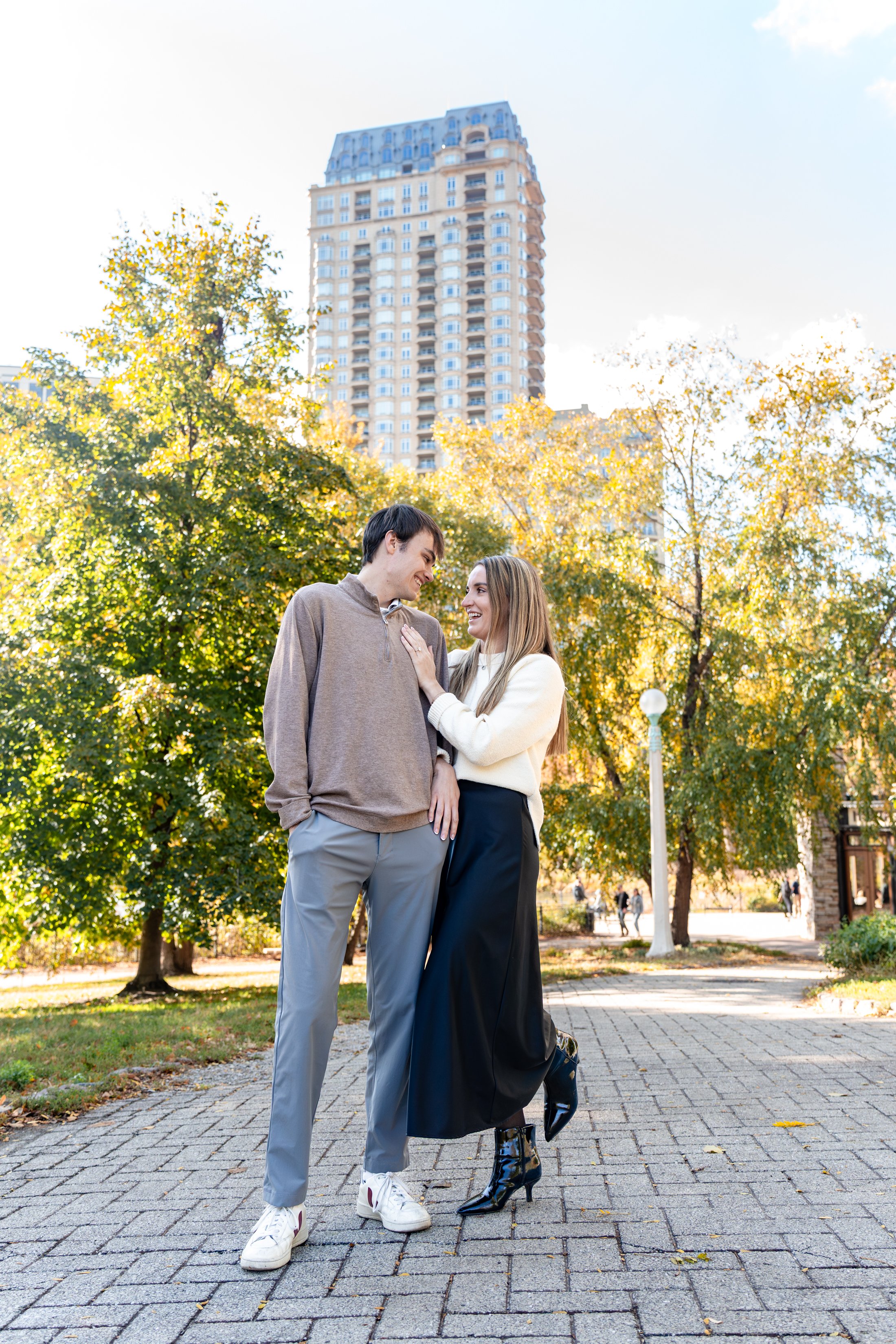 A young couple standing together on a paved pathway in a park, smiling and looking into each other's eyes, surrounded by green trees with a tall building in the background on a sunny day.