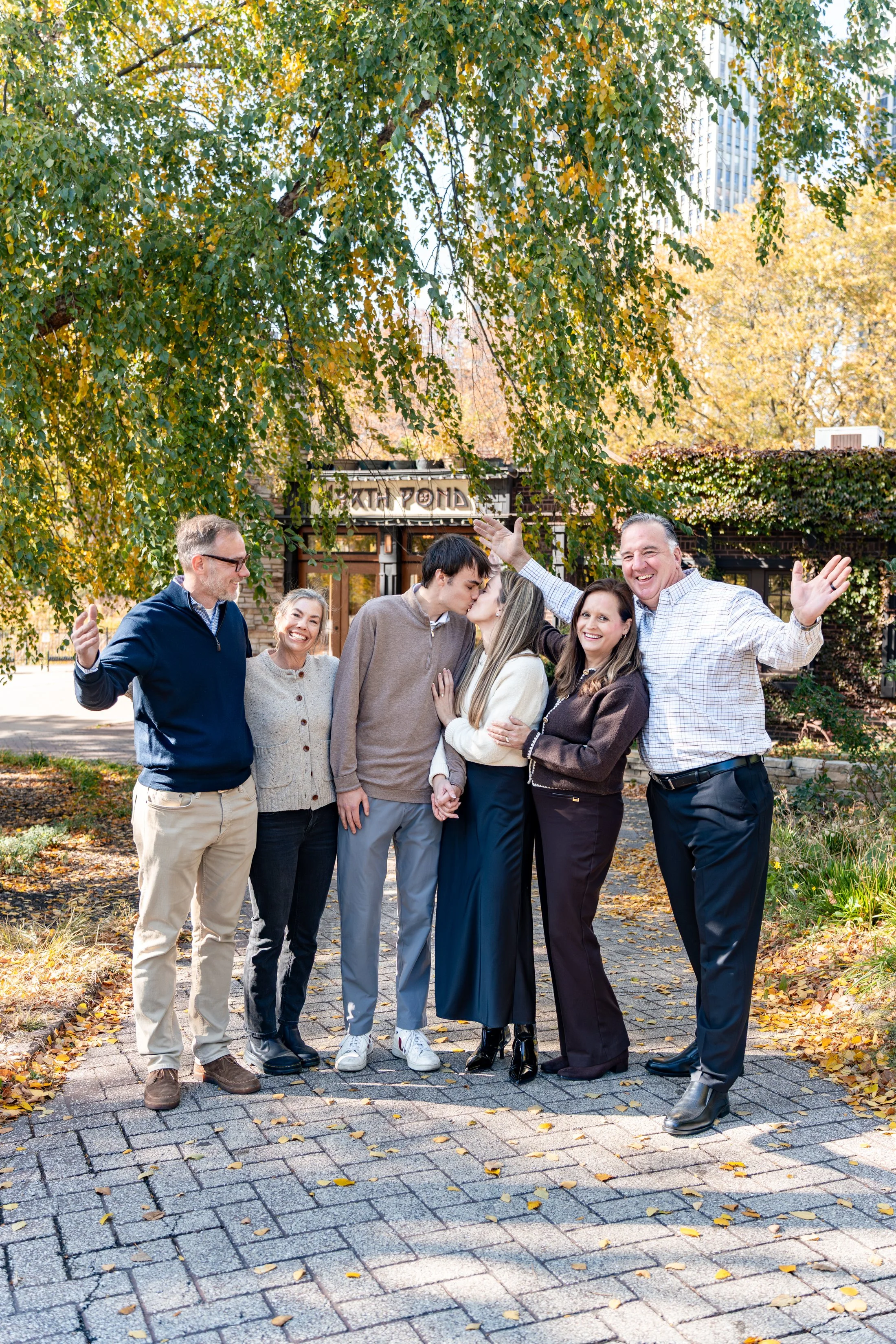 A group of six people, three men and three women, standing outdoors on a stone path, celebrating and smiling under a large green tree in a park during fall. Two people are kissing as others cheer.