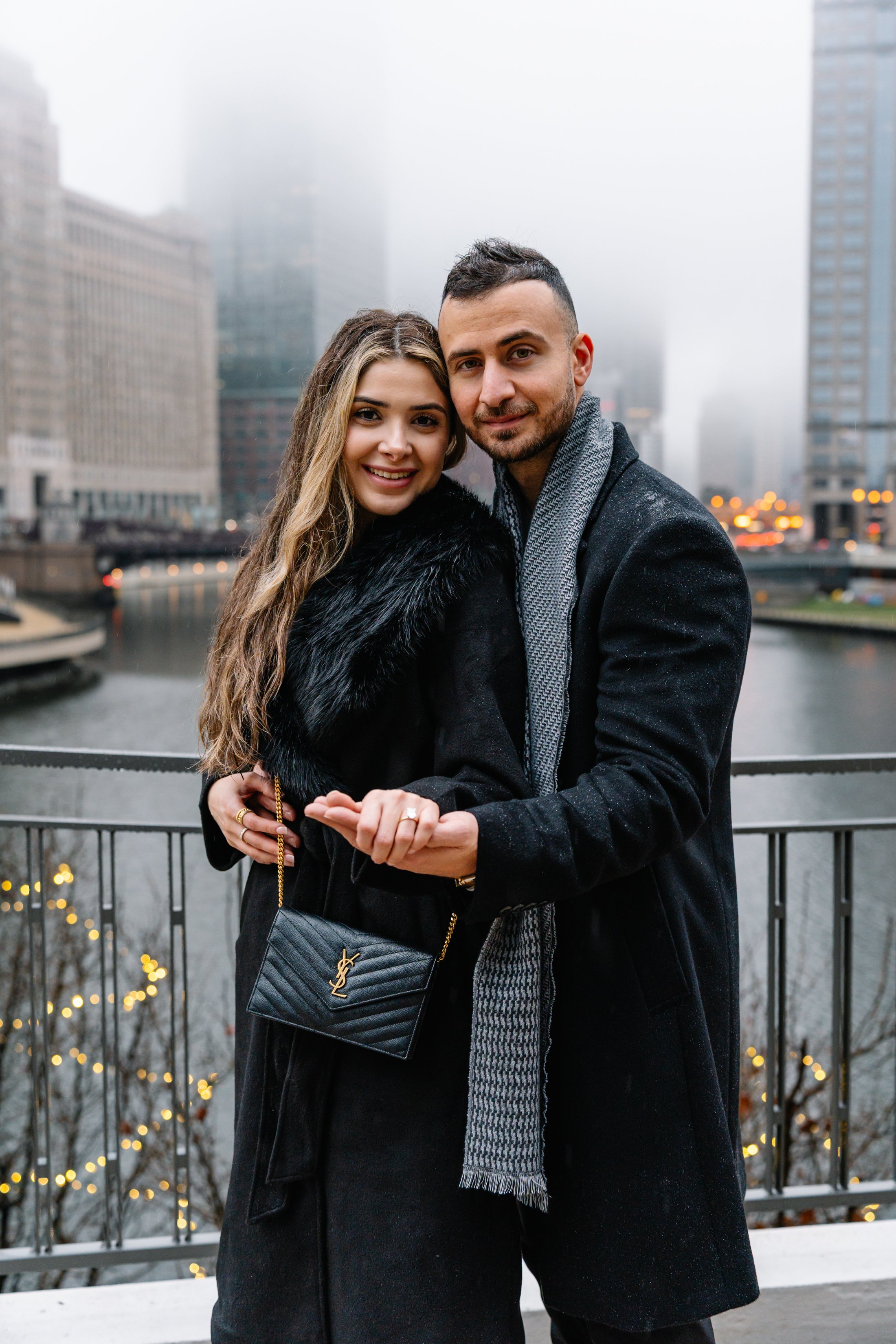 A smiling couple standing close together on a balcony overlooking a city canal with foggy skyscrapers in the background. The woman has long wavy hair, wearing a black coat with a fur collar and carries a black Yves Saint Laurent purse. The man has sh