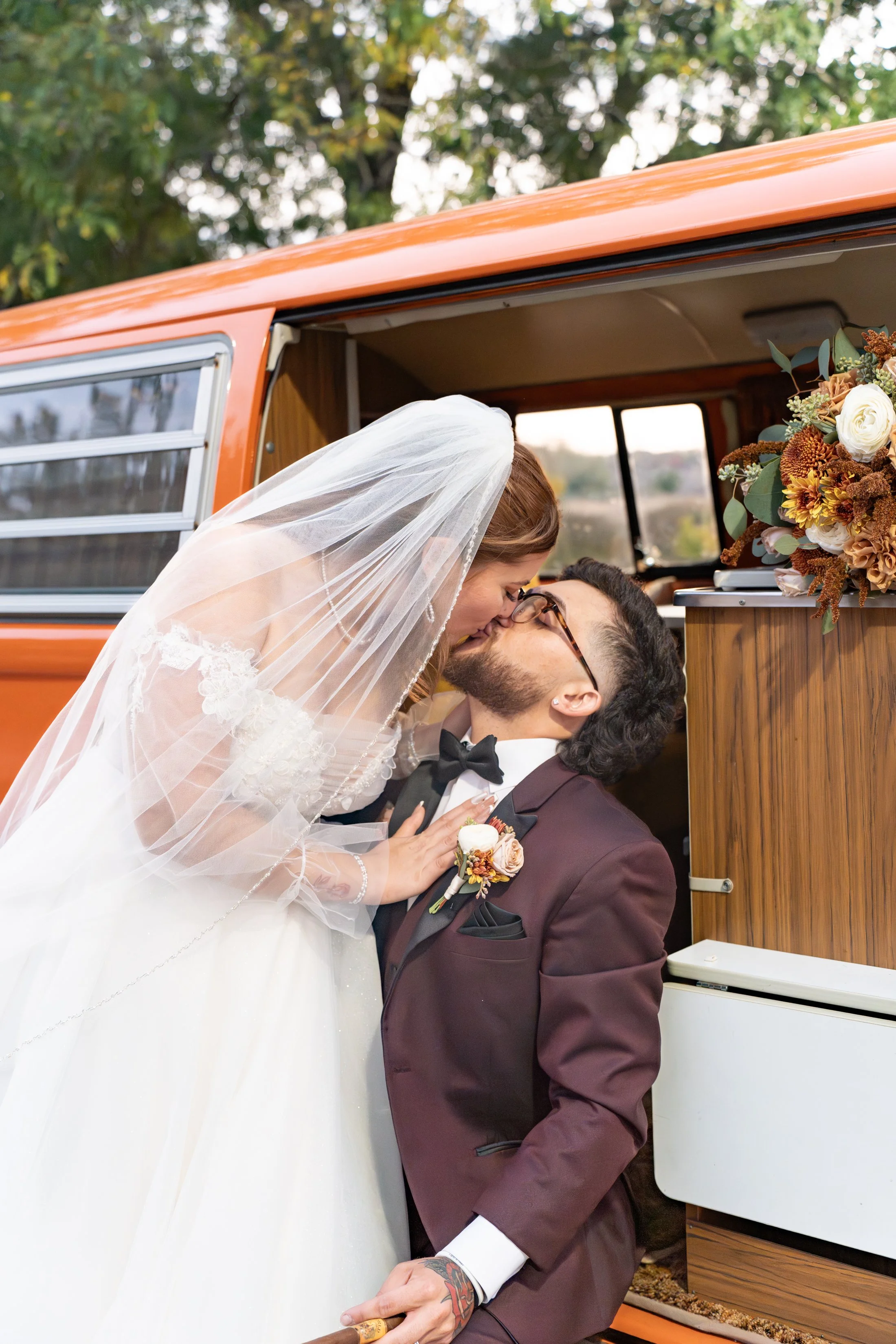 A bride and groom sharing a kiss outside a vintage orange camper, with the bride leaning over the groom, dressed in a white wedding gown and veil, and the groom in a burgundy suit with a boutonniere and bow tie, during a wedding celebration.