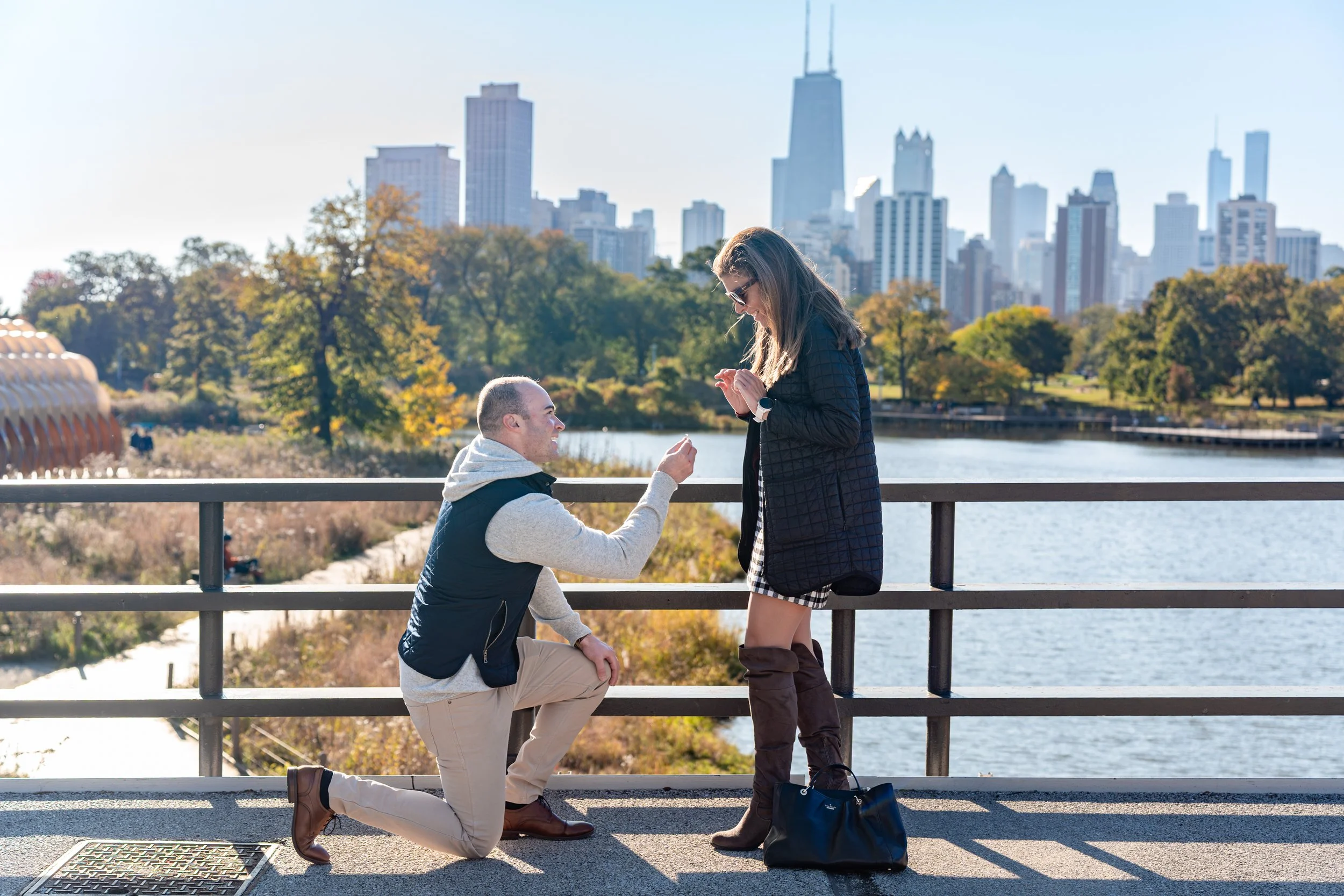 A man proposing to a woman on a bridge over water with city skyline in the background during daytime.