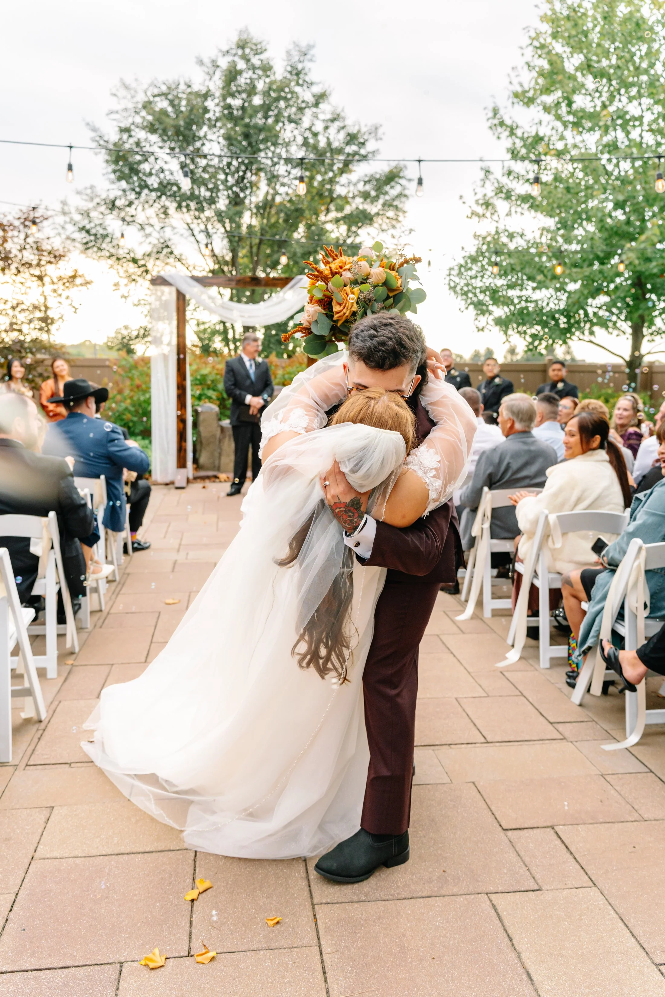 A couple hugging during their wedding ceremony, with guests seated in chairs and a decorated arch in the background outdoors.