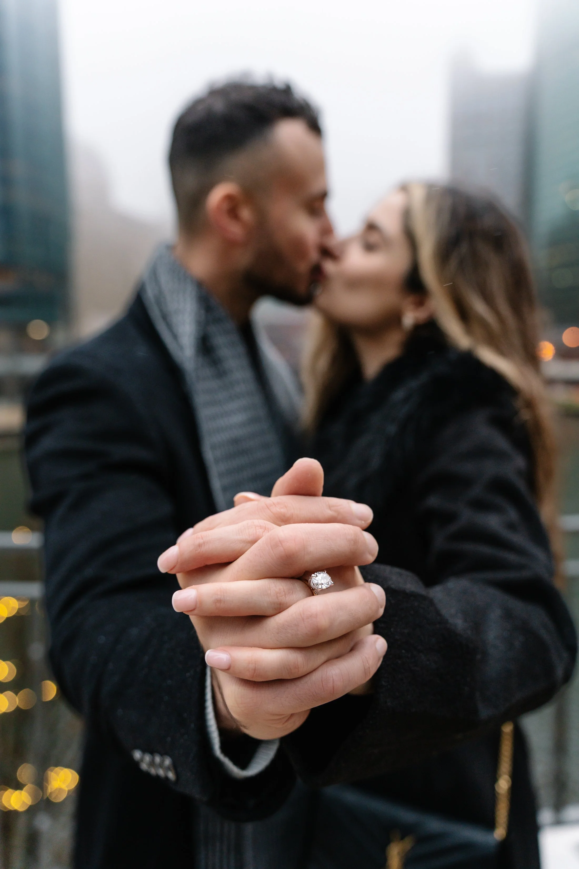 A couple kissing in an urban setting, with the woman showing her hand with an engagement ring in focus.