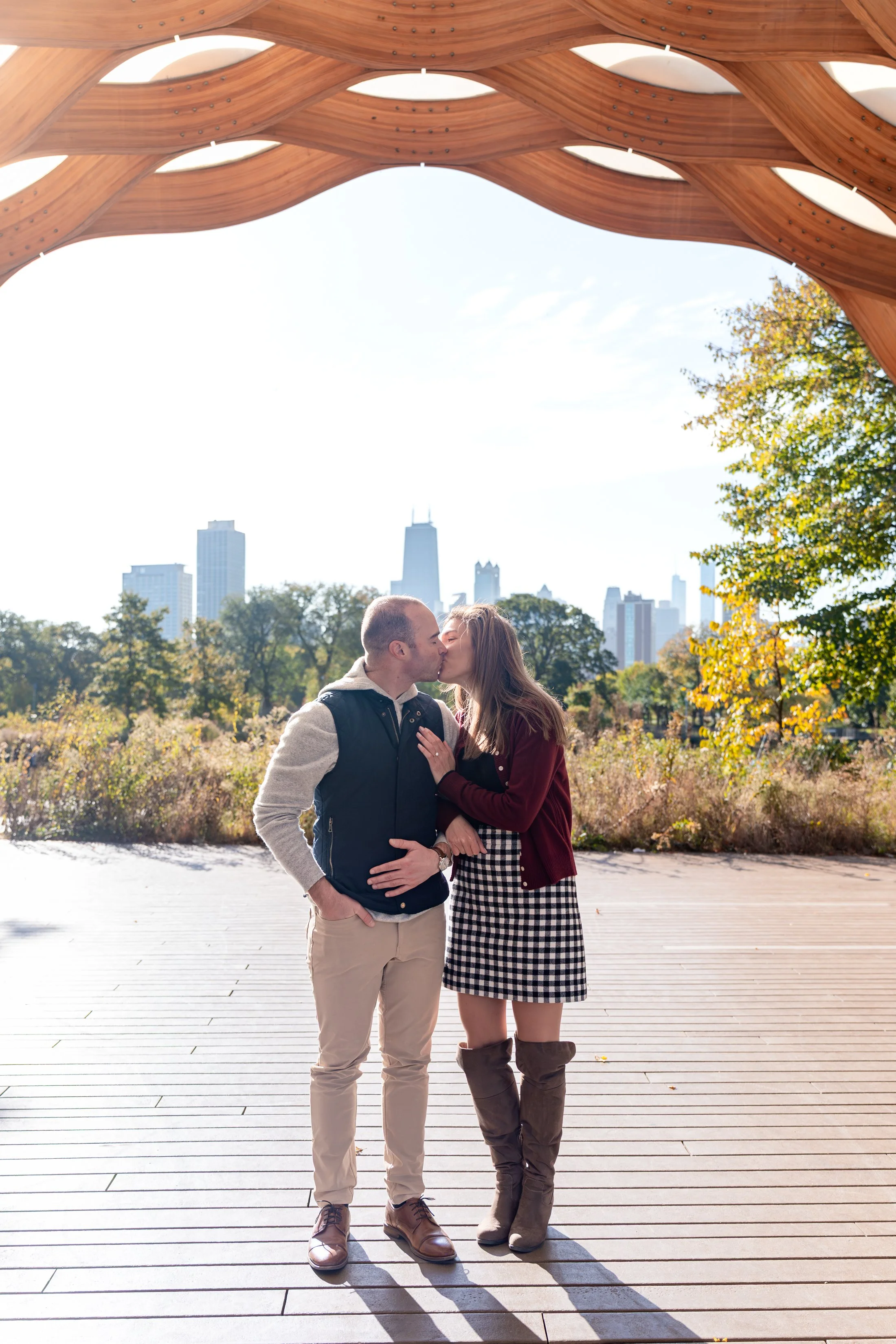 A couple kissing outdoors under a wooden canopy with a city skyline in the background on a sunny day.