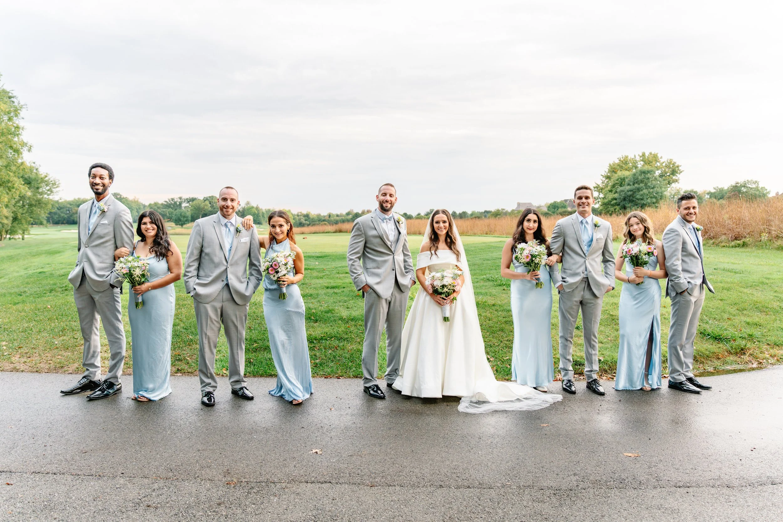 A wedding party standing outdoors on a paved path, with a green field and trees in the background, smiling for the camera.