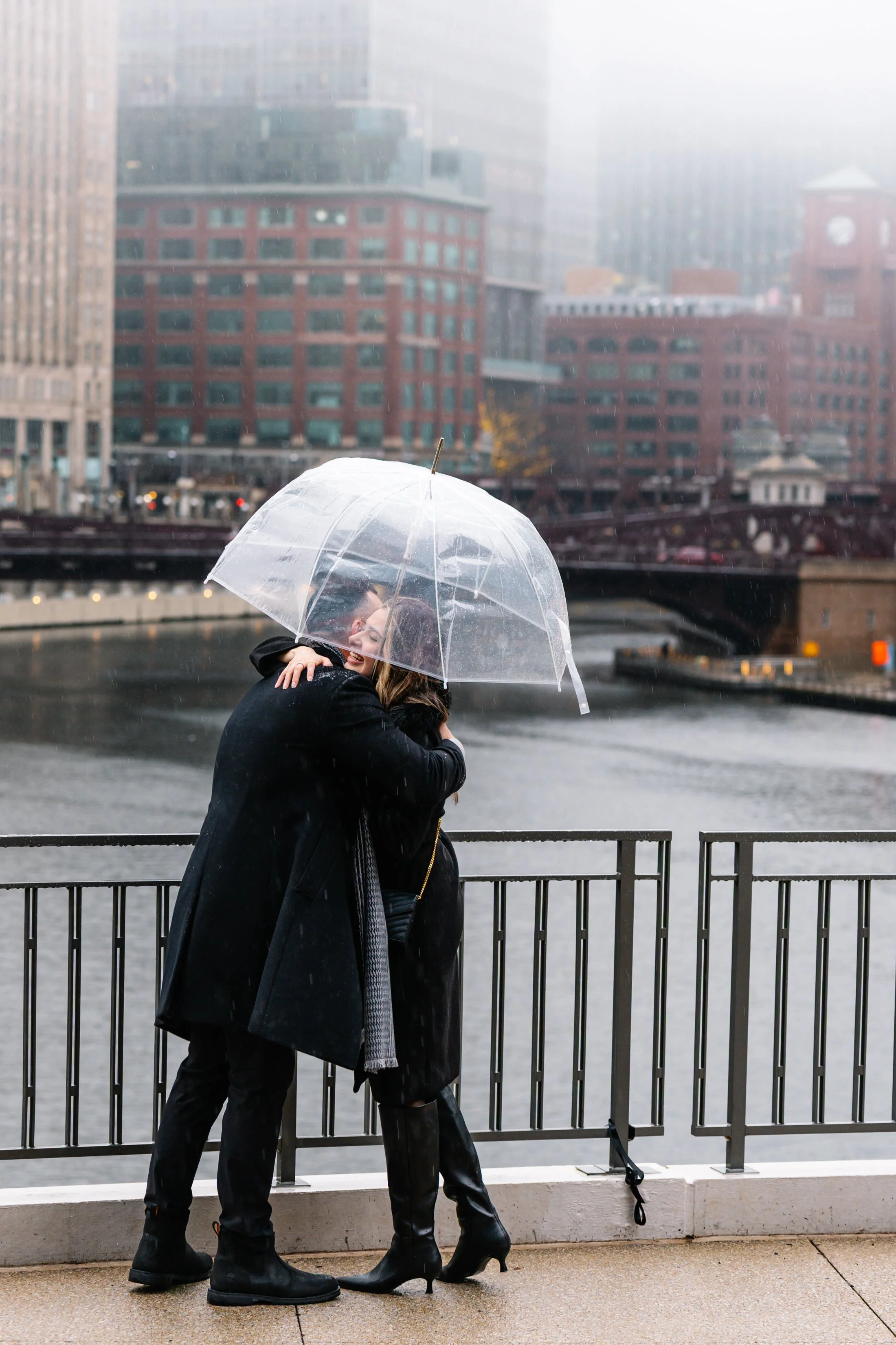 A couple embraces under a transparent umbrella on a rainy day by a river, with city buildings in the background.