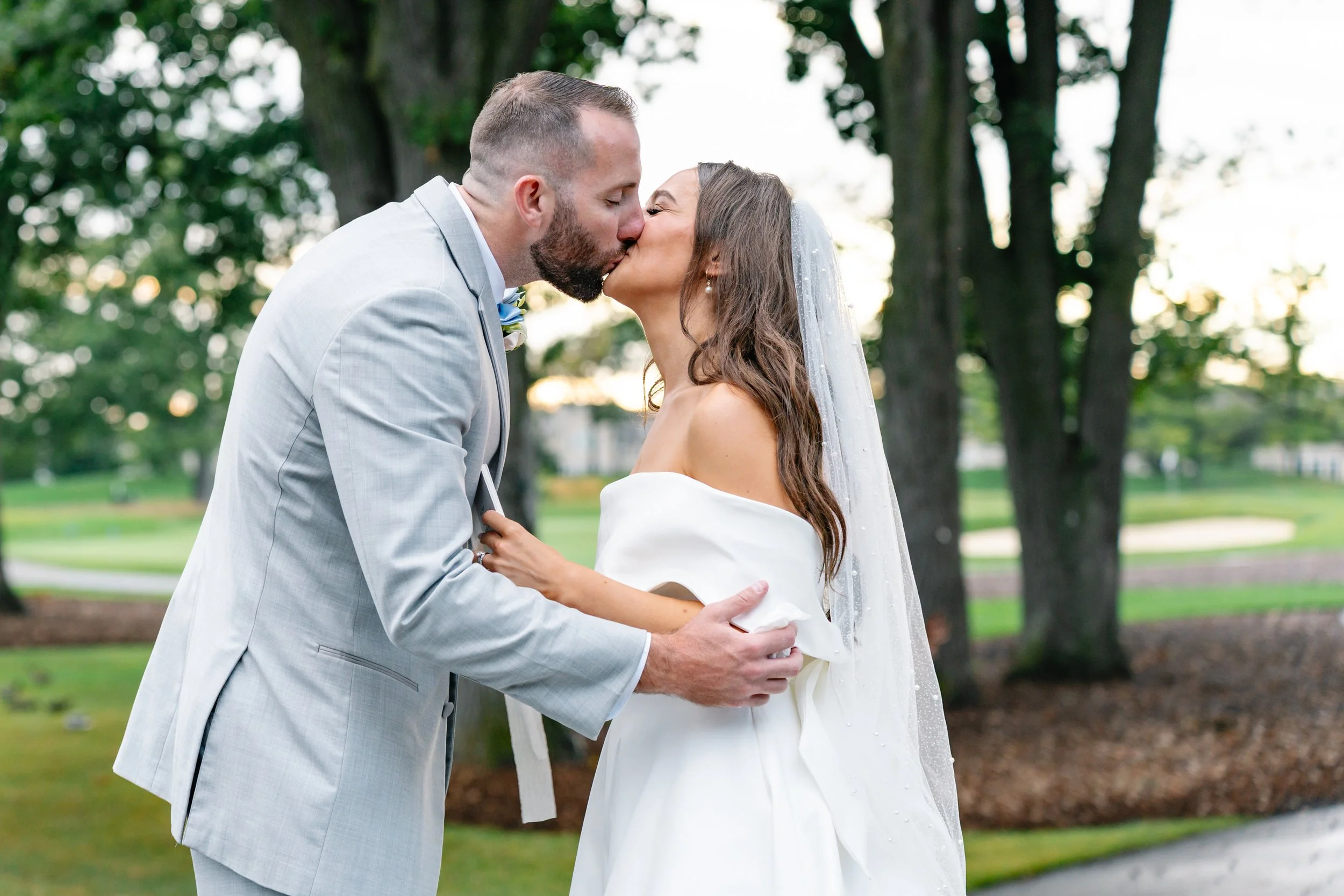 A bride and groom kissing outdoors in a park during their wedding, surrounded by trees.