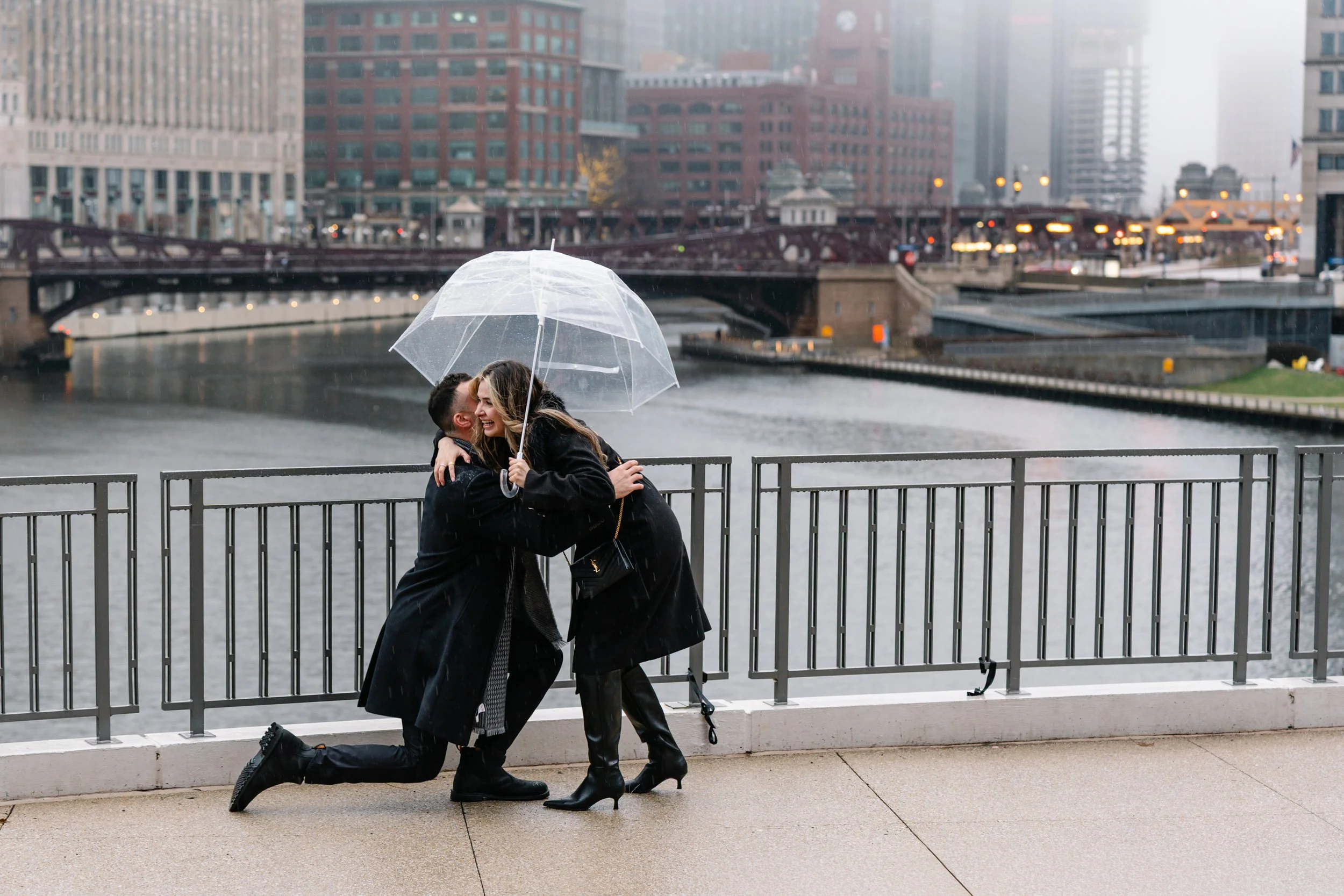 A couple hugging and smiling under an umbrella on a rainy day by a river in a city, with tall buildings and a bridge in the background.