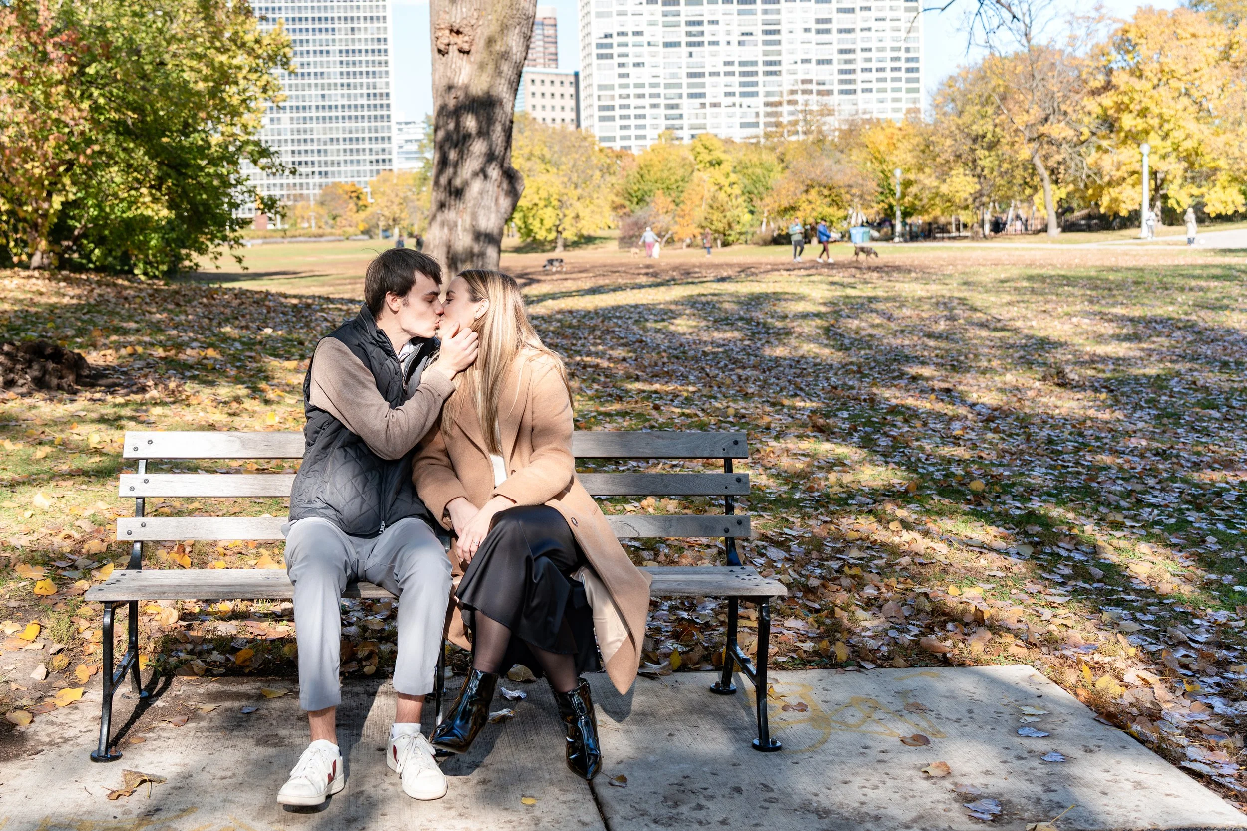 A young couple sitting on a park bench kissing, autumn leaves on the ground, city buildings in the background.