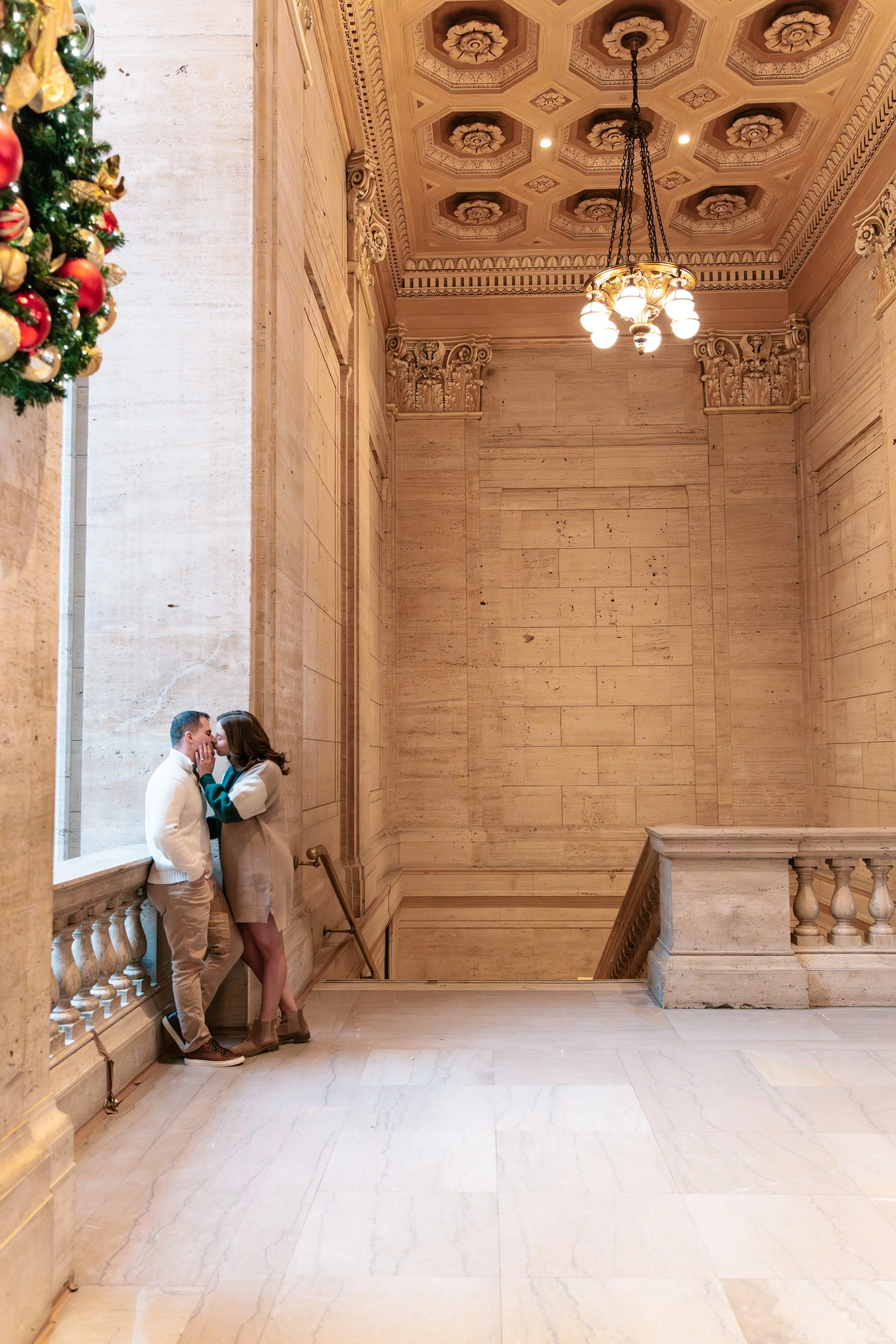 A man and woman sharing a kiss in a grand, historic marble building with intricate ceilings, a vintage chandelier, and a Christmas wreath partially visible on the left side.