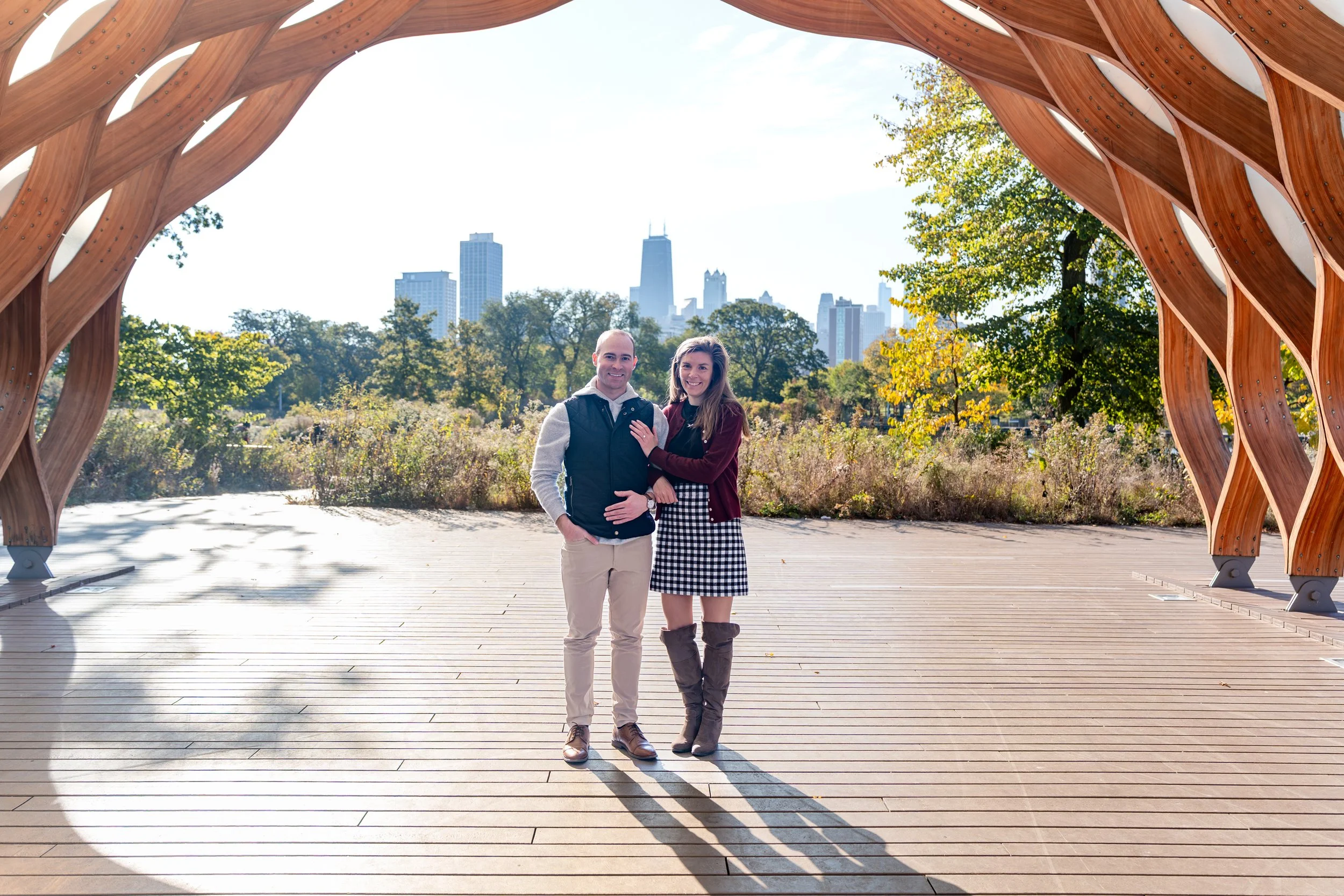 A couple standing under a large wooden arch in a park with trees and city skyline in the background, bright sunlight overhead.